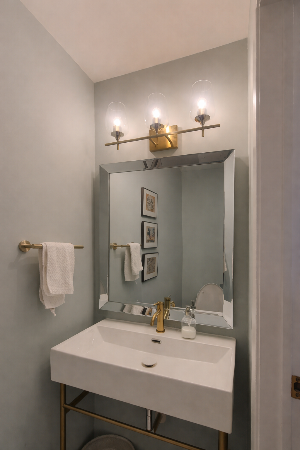 A bathroom renovation with a white sink, gold faucet, large mirror, wall art, towel rack with white towels, a three-light vanity fixture above the mirror, and a soap dispenser on the sink.