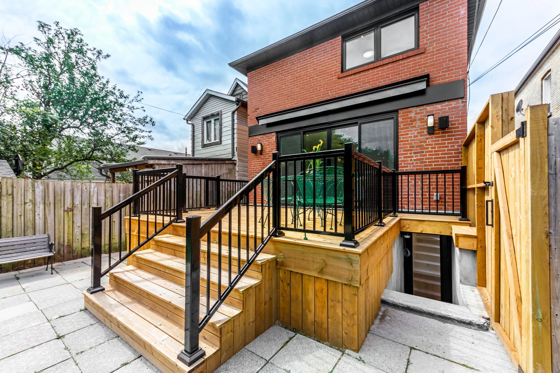 Backyard patio with a new wooden deck and black metal railing, steps leading down to a paved area, and a house with a brick exterior. There is a table and chairs on the deck, and a wooden privacy fence.