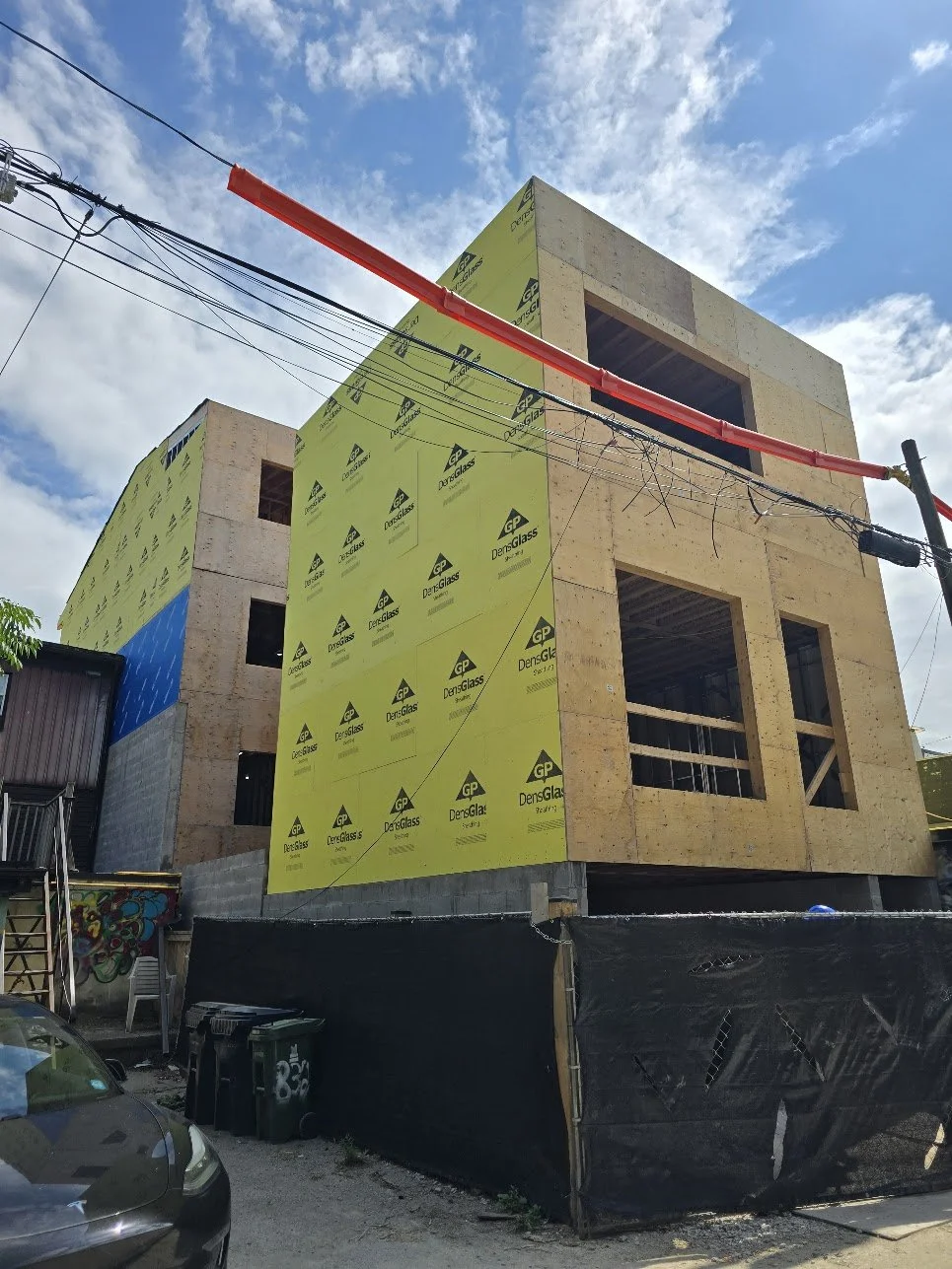 Construction site of a multi-story building with wooden framing and yellow insulation panels, black construction fencing, parked black car, utility wires, and partly cloudy sky.