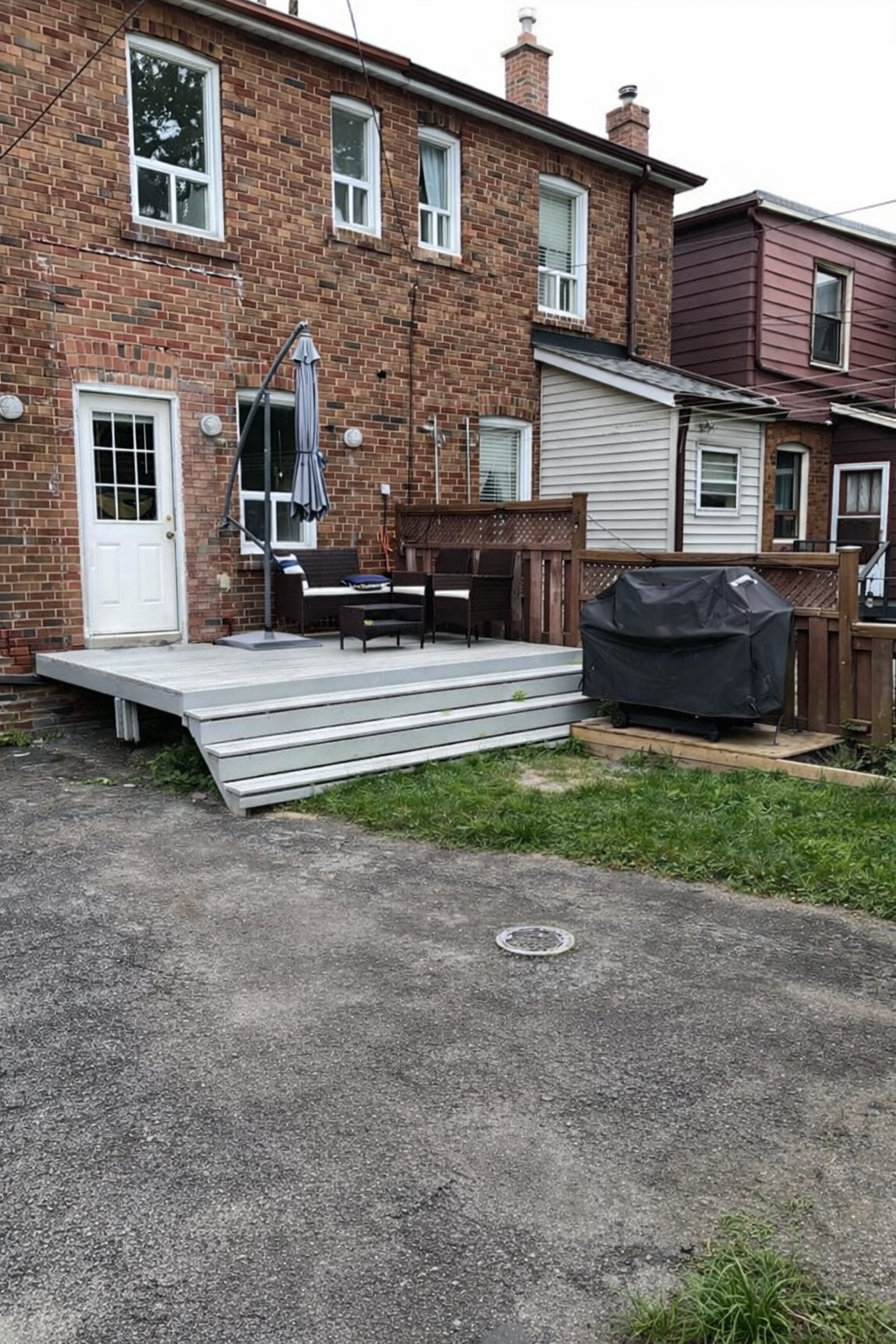 Backyard patio with a small set of wooden stairs leading to a wooden deck. The deck has outdoor furniture, including a table with an umbrella and a grill covered with a black cover. The deck is attached to a brick house with multiple windows and a white door. There is a gravel area in the foreground and neighboring houses visible in the background.