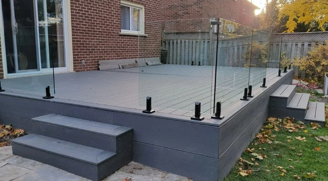 Backyard composite deck with glass railing, steps leading up to it, and brick house in background, surrounded by autumn leaves.
