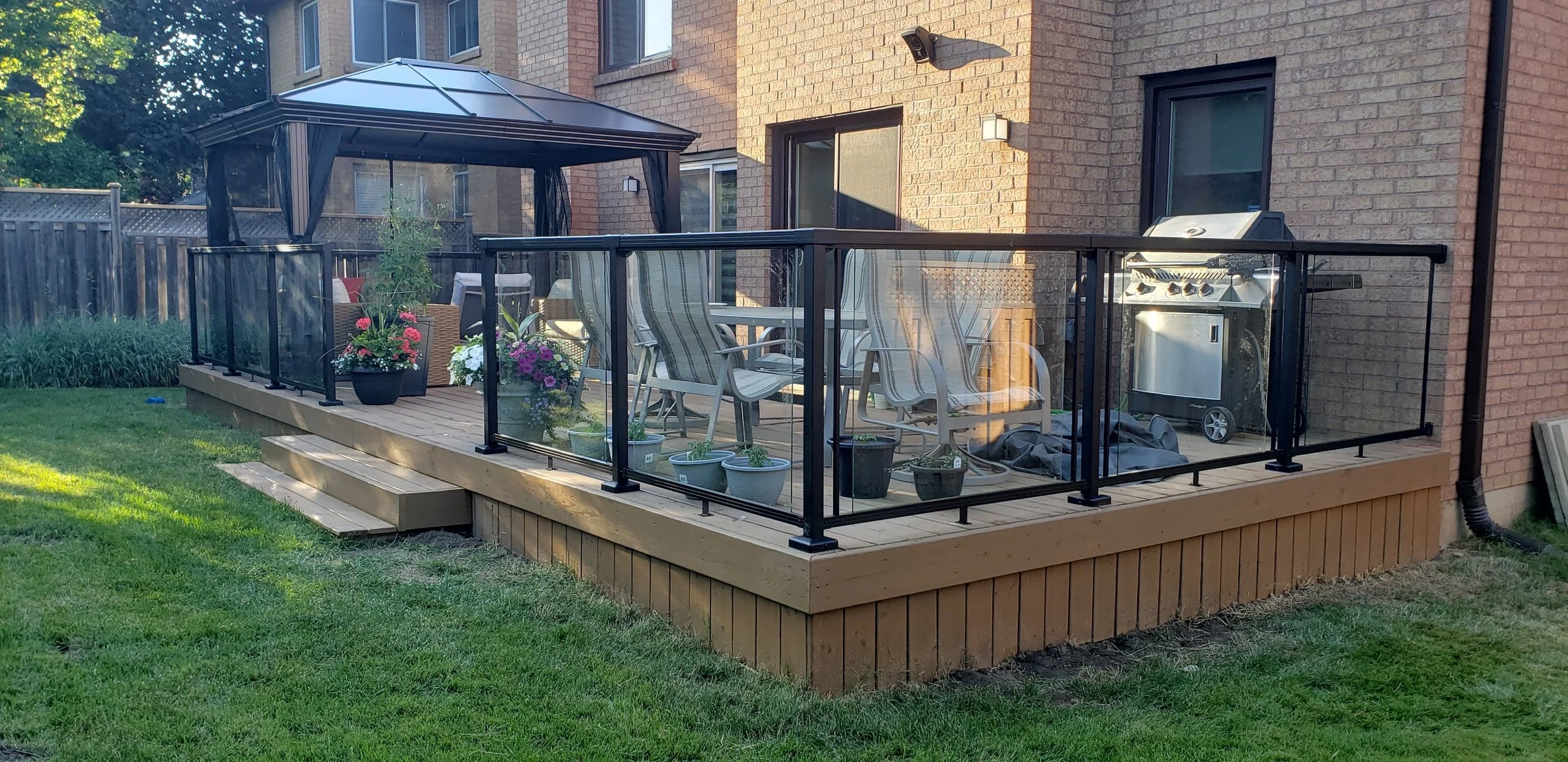 A backyard deck with black metal railing, decorated with potted plants and flowers, furnished with a table and chairs, a grill, and a gazebo in the background, attached to a brick house.