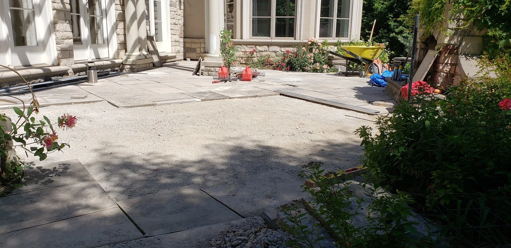 A backyard patio area under construction featuring partially installed paving stones, flowers, and gardening tools, with a house with large windows in the background.