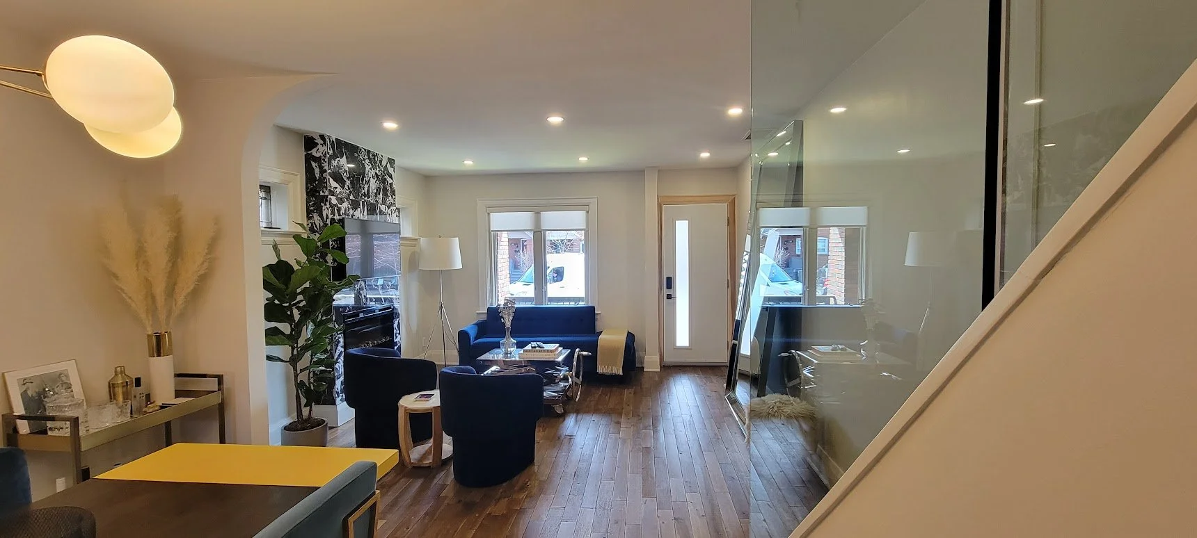 Living room with wooden floor, blue sofa, black armchairs, coffee table, fireplace with black and white patterned wallpaper, large window, standing lamp, greenery, and modern decor, separated by glass wall on the right.
