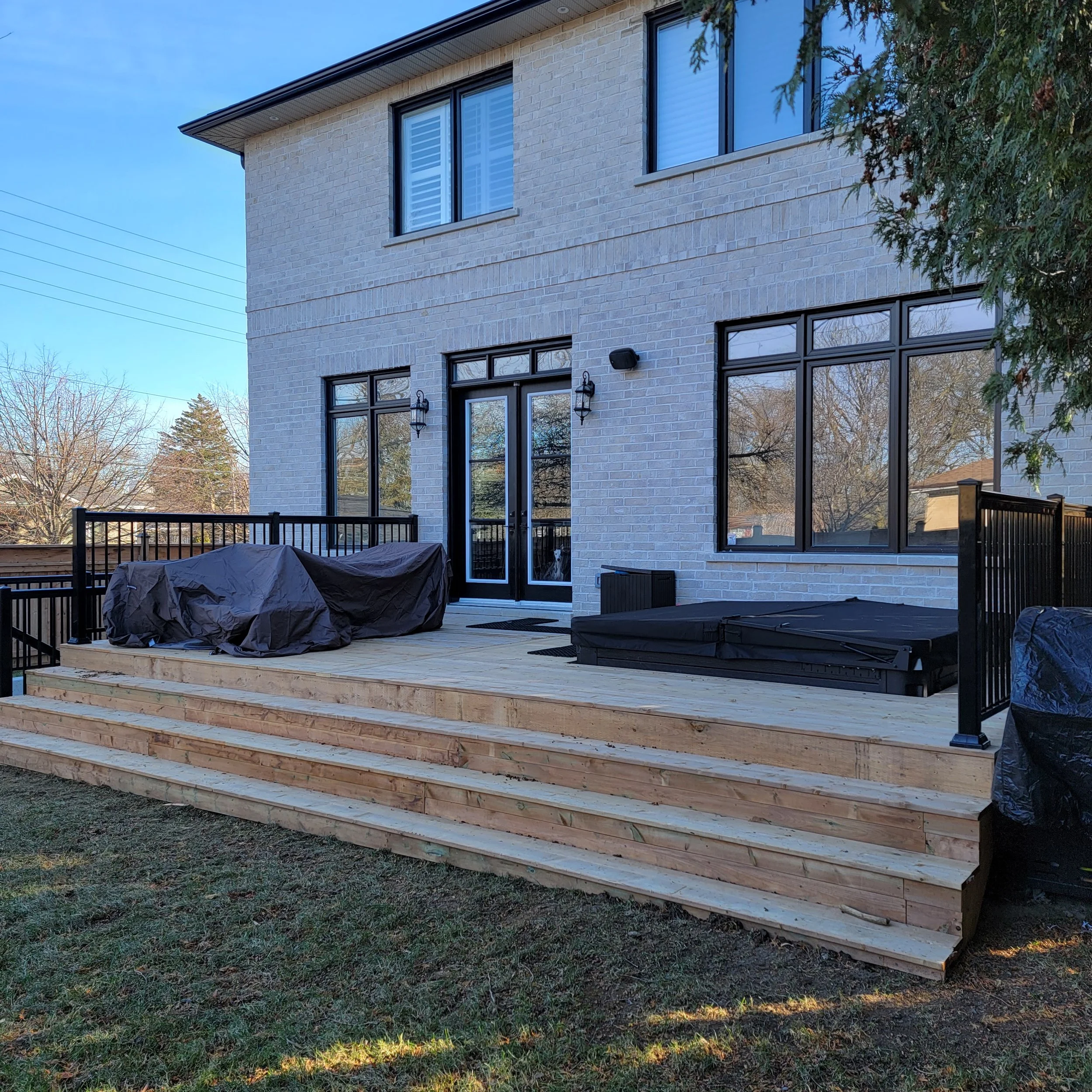 A newly built wooden deck attached to the back of a white brick house, with stairs leading down to a grassy yard. The deck has black metal railings, a covered grill, a seating area with cushions, and is illuminated by outdoor wall lights.
