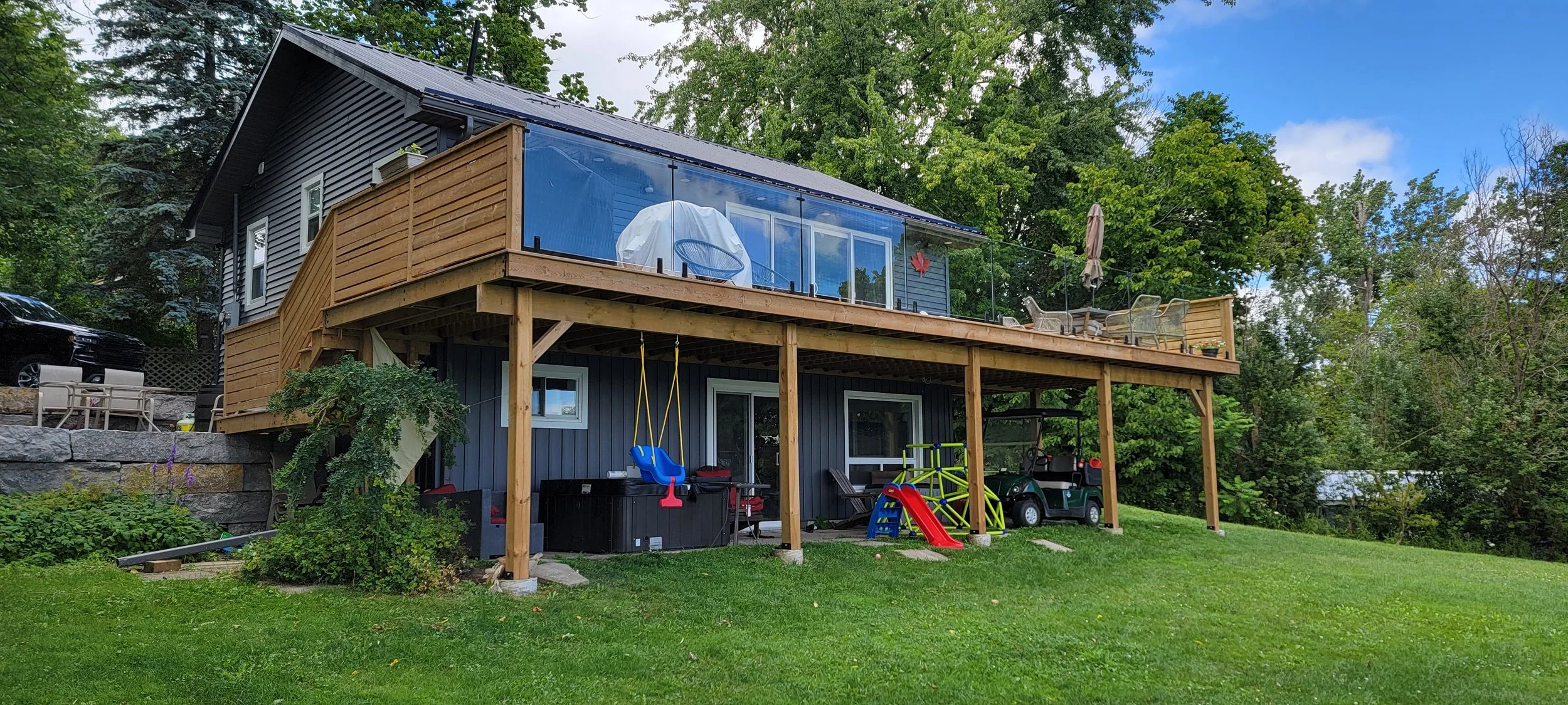 Two-story house with a large wooden deck, enclosed balcony on the upper level, and a grassy yard underneath. The yard has children's play equipment, a golf cart, and outdoor furniture. The house is surrounded by trees and greenery.