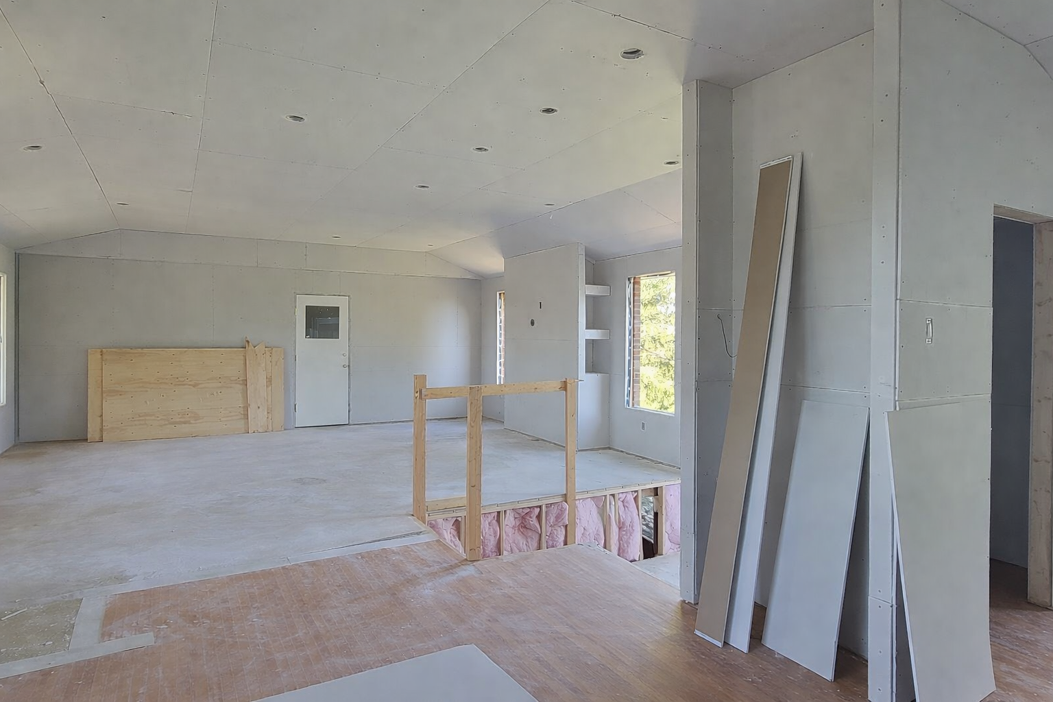 Interior of a house under construction with drywall, wooden framing, and unfinished flooring, with windows and construction materials visible.