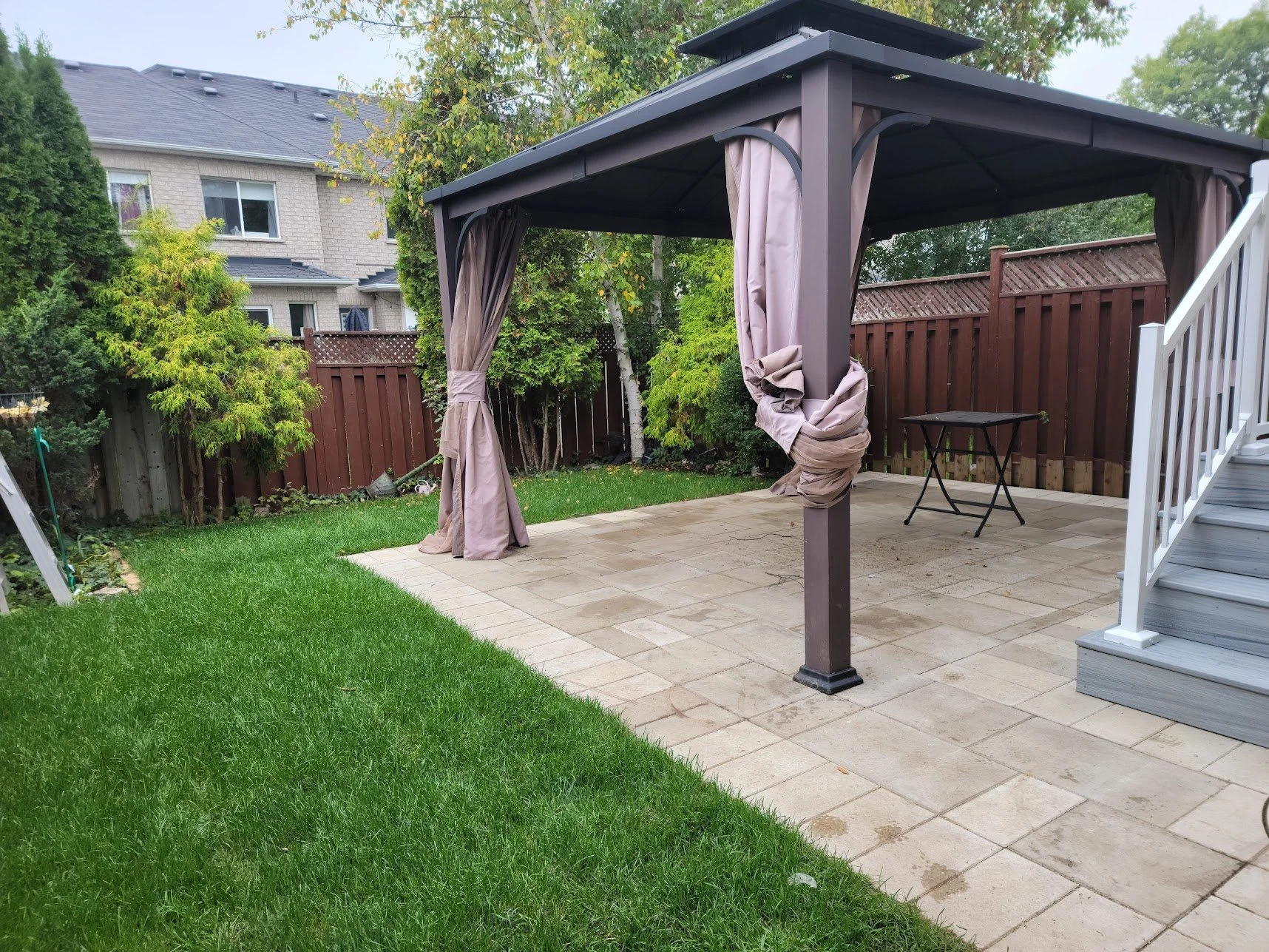 Backyard patio with a covered gazebo, brown curtains, and a small black table. Surrounding greenery includes a lawn and trees, with a house visible in the background.