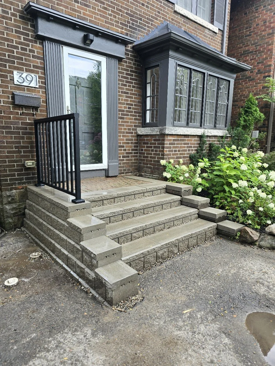 Brick house with a glass front door, black metal railing, and stone steps leading to the entrance; white hydrangea bushes beside the steps.