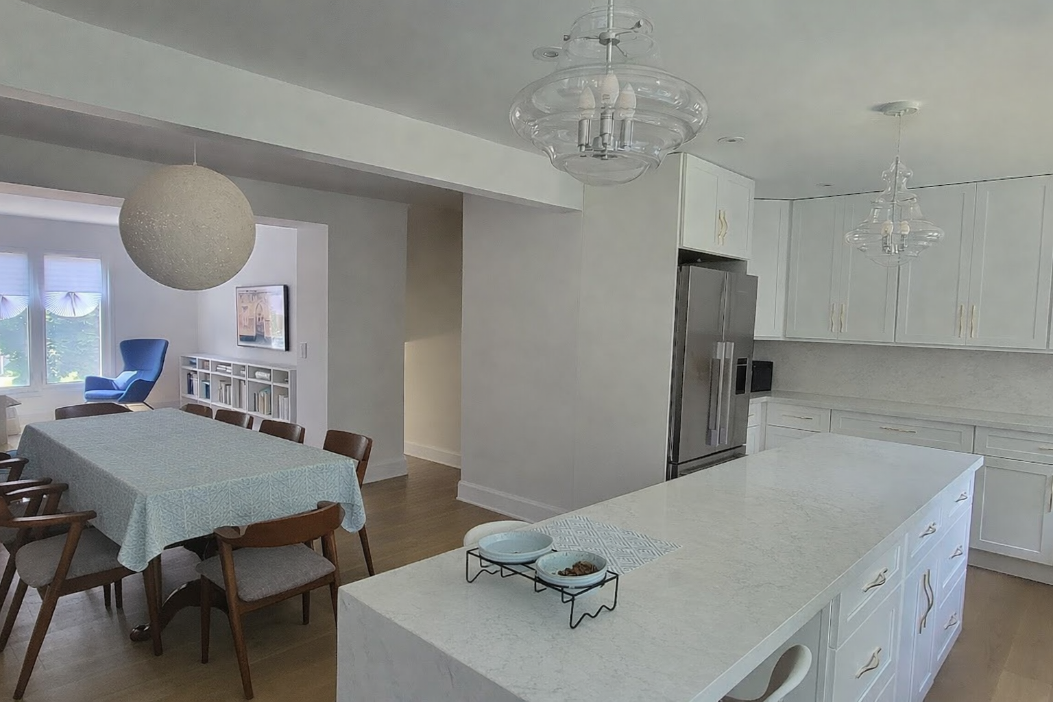 Modern kitchen and dining area with white cabinets, island countertop, stainless steel refrigerator, and a dining table with wooden chairs. Light fixtures hang from the ceiling.