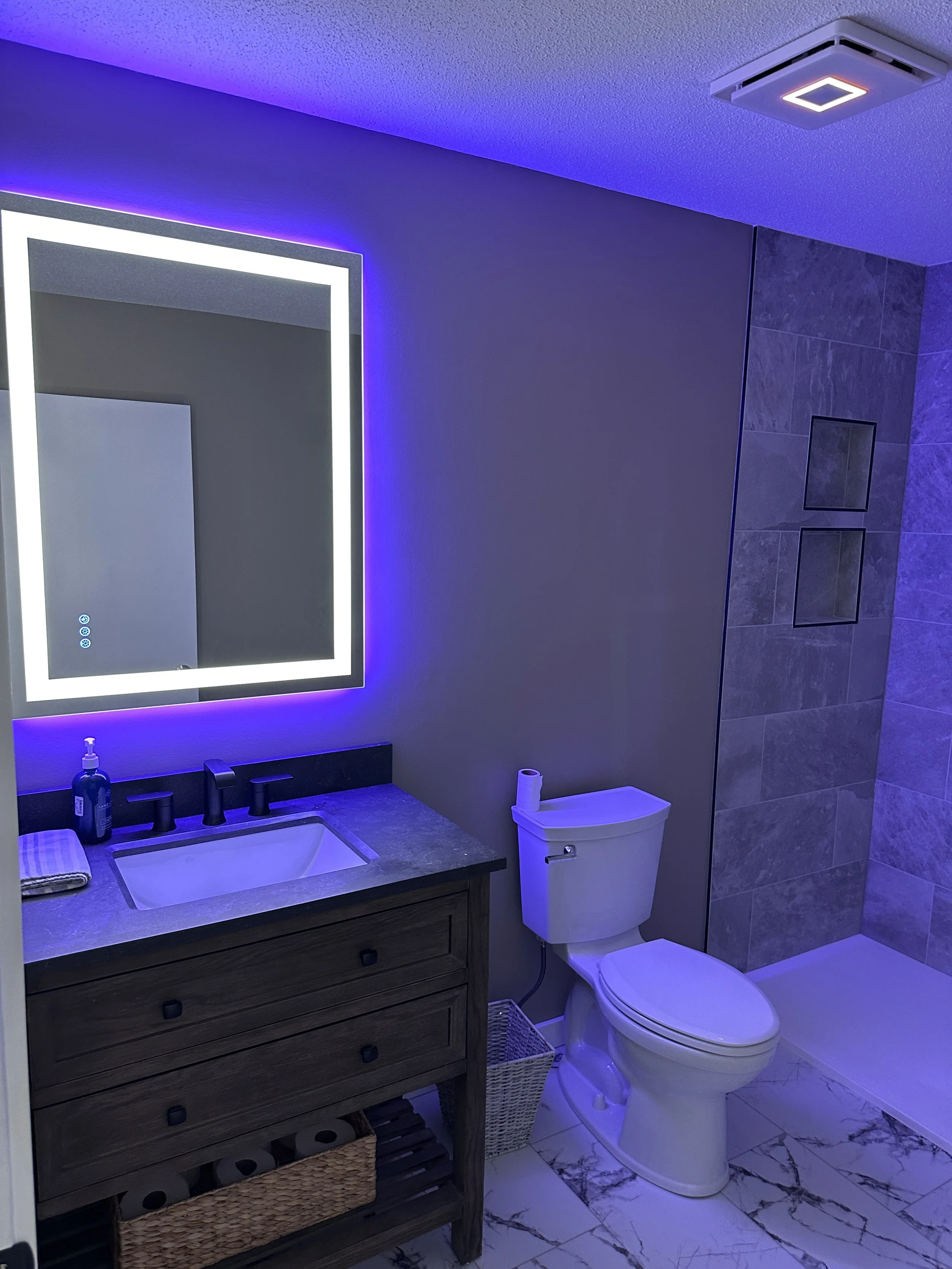 Modern bathroom with a illuminated mirror, sink with black faucet, toilet with roll of toilet paper on top, and a shower area with gray tiles. The purple LED lighting highlights the space.