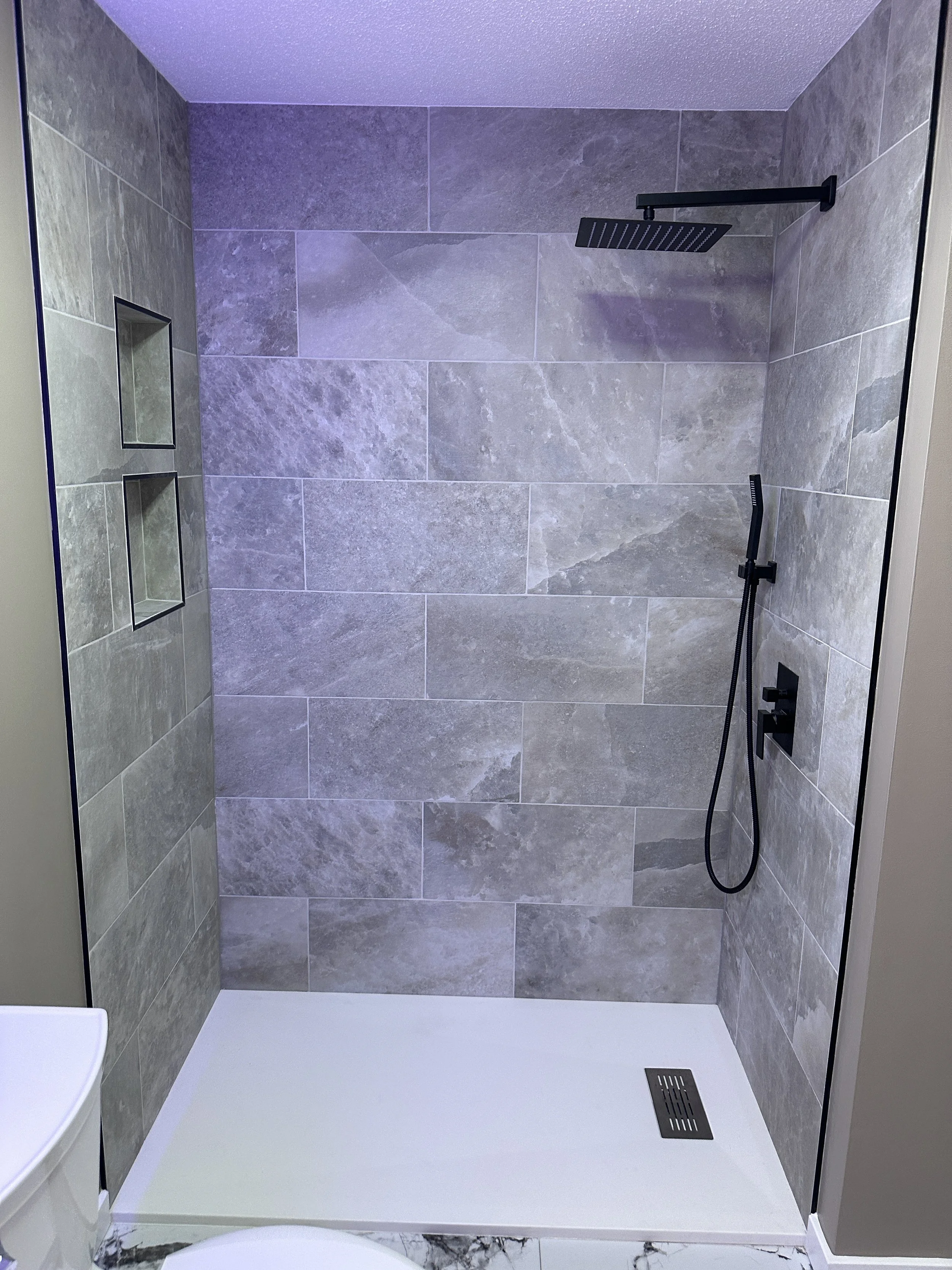 A modern walk-in shower with large grey stone tiles, black fixtures, a rainfall showerhead, a handheld shower spray, two recessed shelves, and a square drain on the white shower floor.