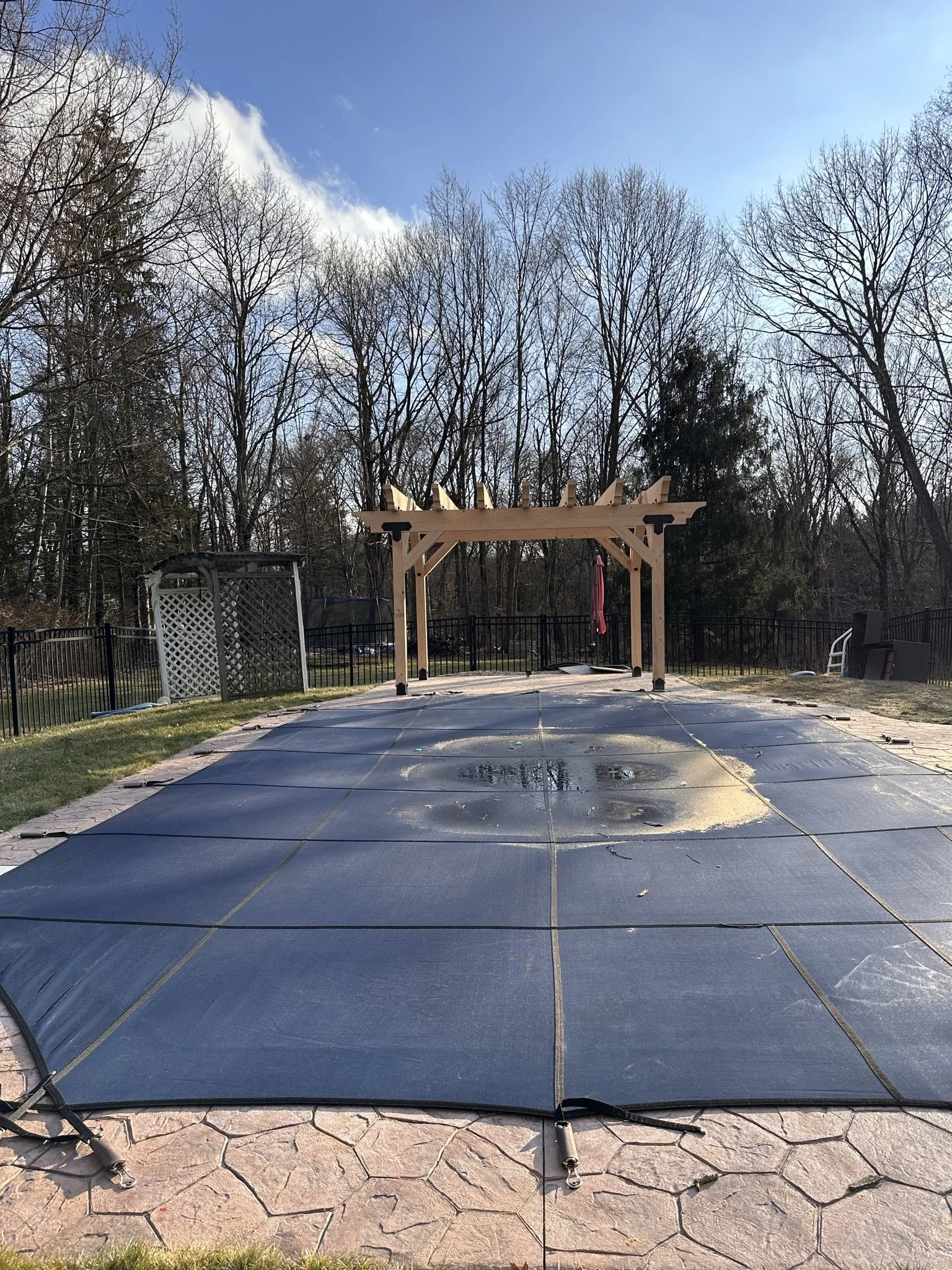 A swimming pool covered with a safety cover, with a wooden pergola at the end, in a backyard surrounded by leafless trees.