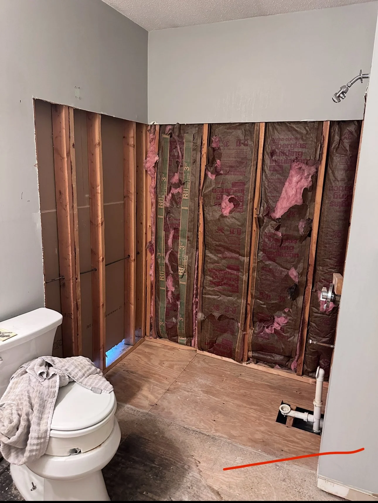 Bathroom with partial wall demolition showing exposed studs, insulation, and plumbing, accompanied by a toilet with a checkered towel draped over it.