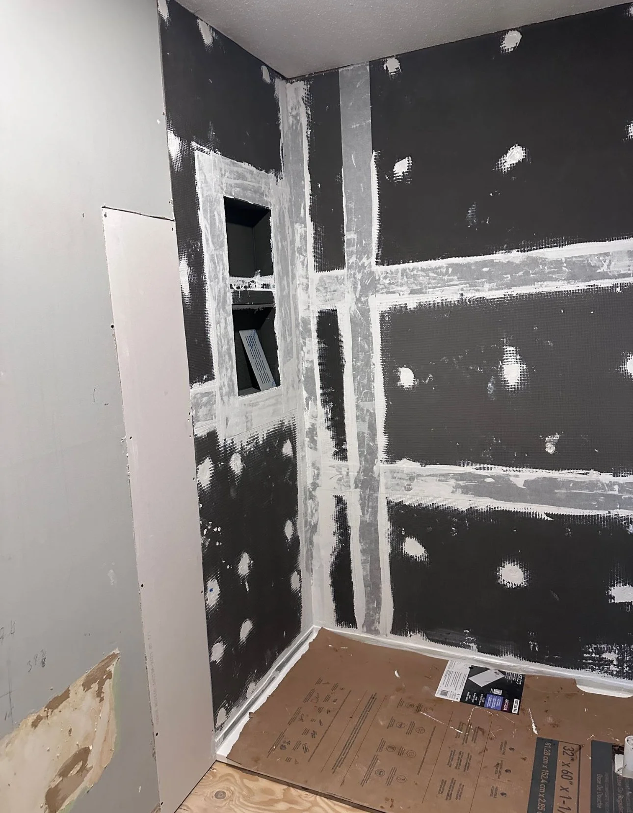 A corner of a room under construction with black drywall, visible bolt holes, an open electrical box, and unfinished drywall patches.