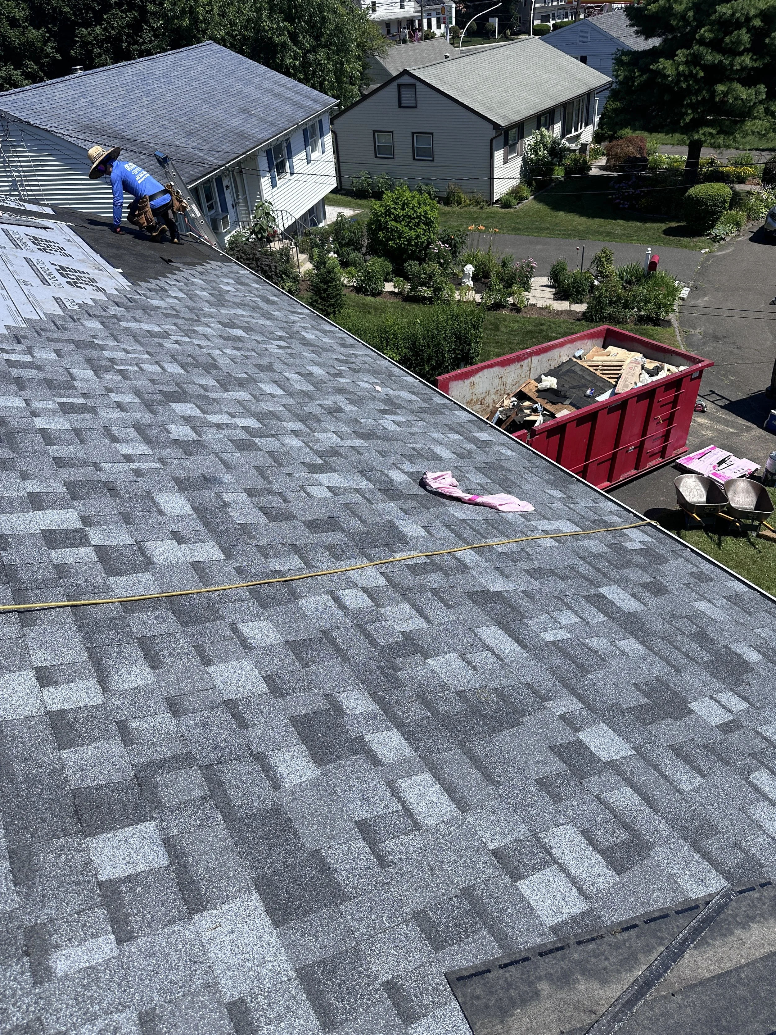 Roof with a worker installing shingles, a utility rope, a cloth, and construction debris in a red dumpster below.