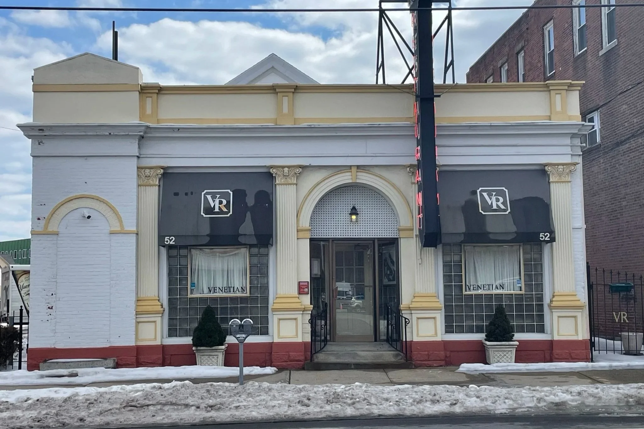 Front of a small building with a sign saying 'VR' and an awning that says 'Venetian.' The building has two large windows with curtains, potted plants on each side of the entrance, and a sidewalk with snow and slush in front. A red vertical sign reads 'RESTAURANT.'