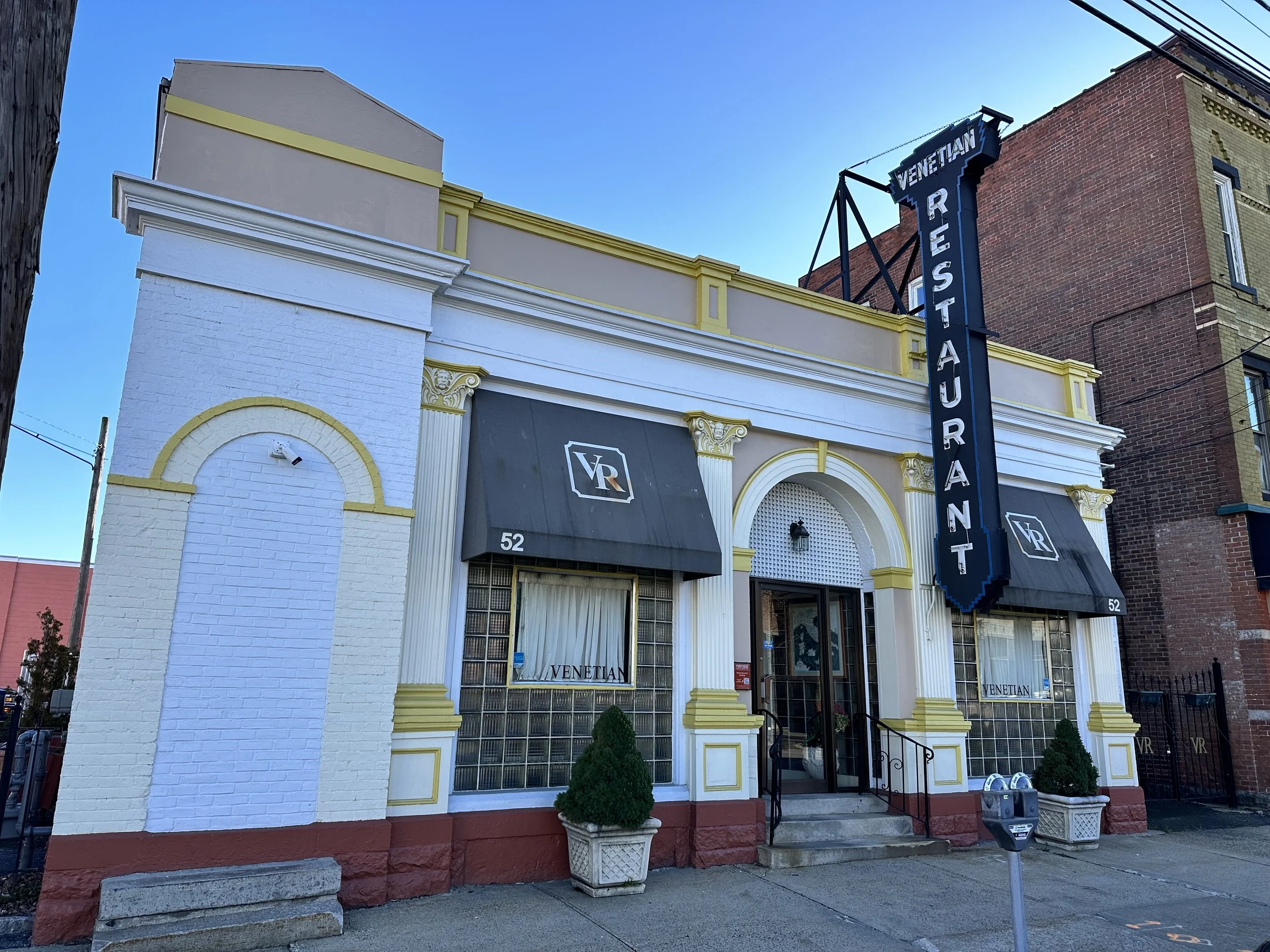 Exterior of a restaurant called Venetian, with a large vertical sign, black awnings with the VR logo, and decorative white and yellow trim. Two potted plants are near the entrance.