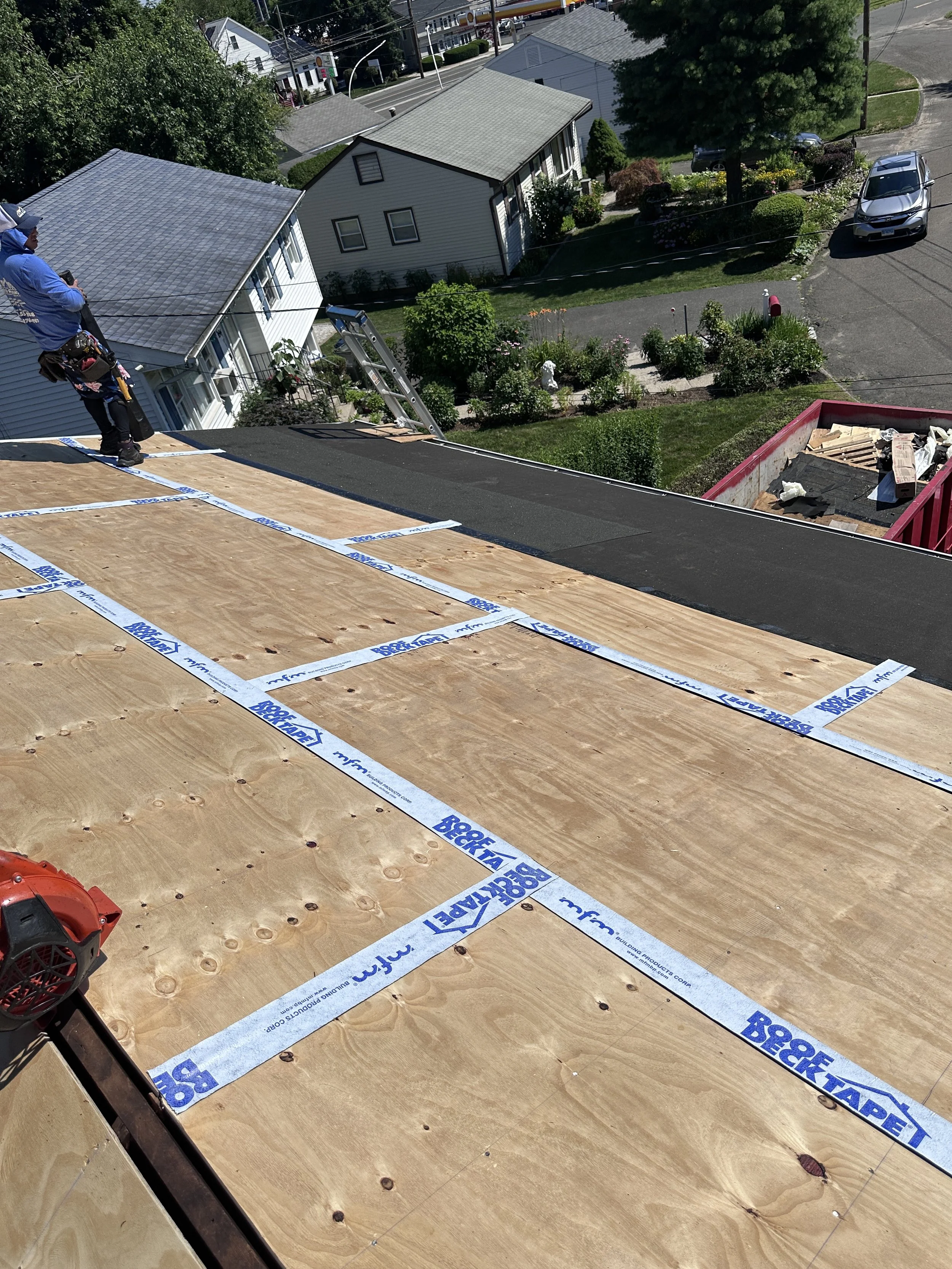 Construction worker standing on a roof under construction, with plywood sheathing and roofing materials, overlooking a residential neighborhood with houses, trees, and a street.
