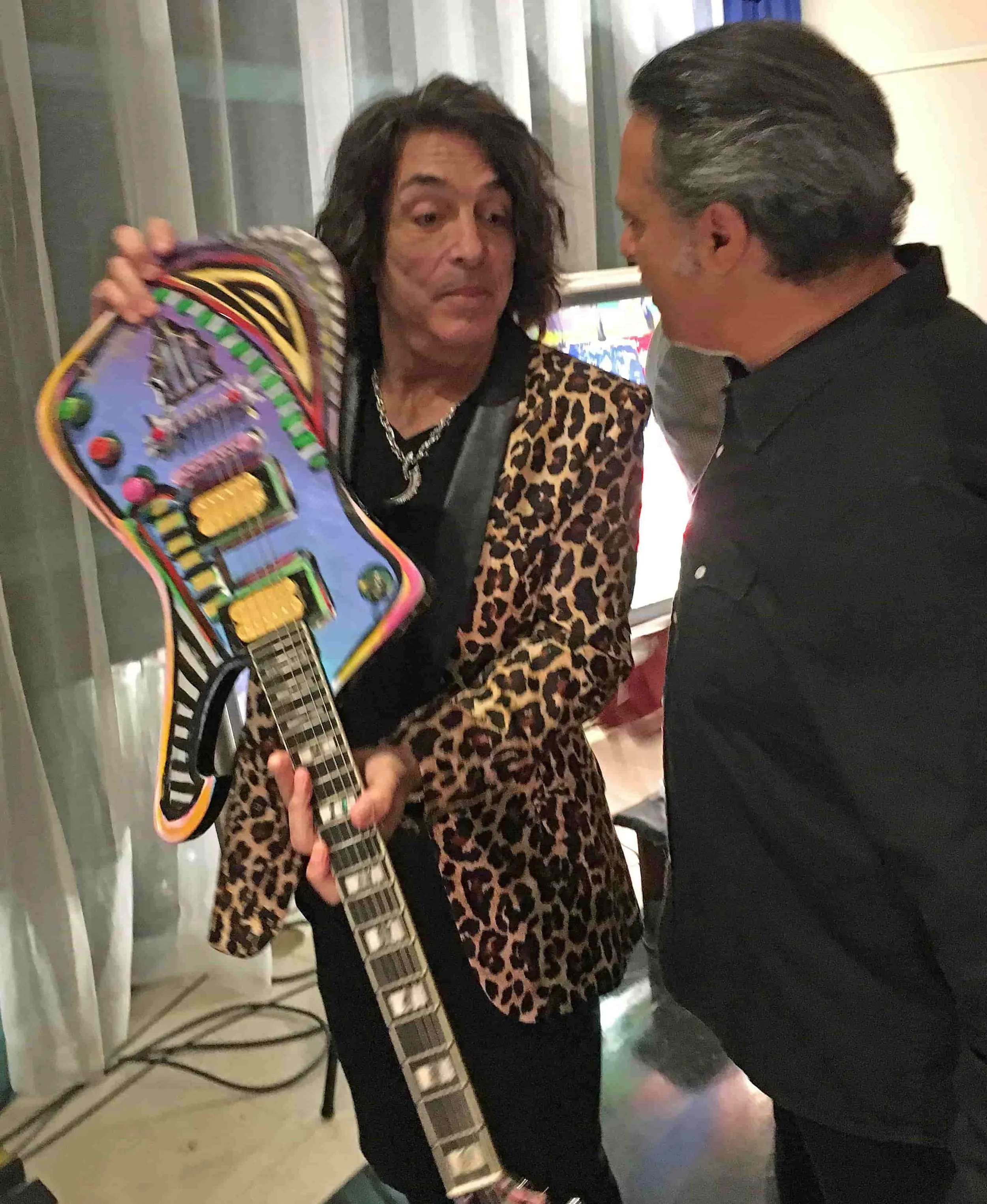 Zarum and KISS band member Paul Stanley, one holding a colorful guitar-shaped sculpture, engaging in conversation indoors.