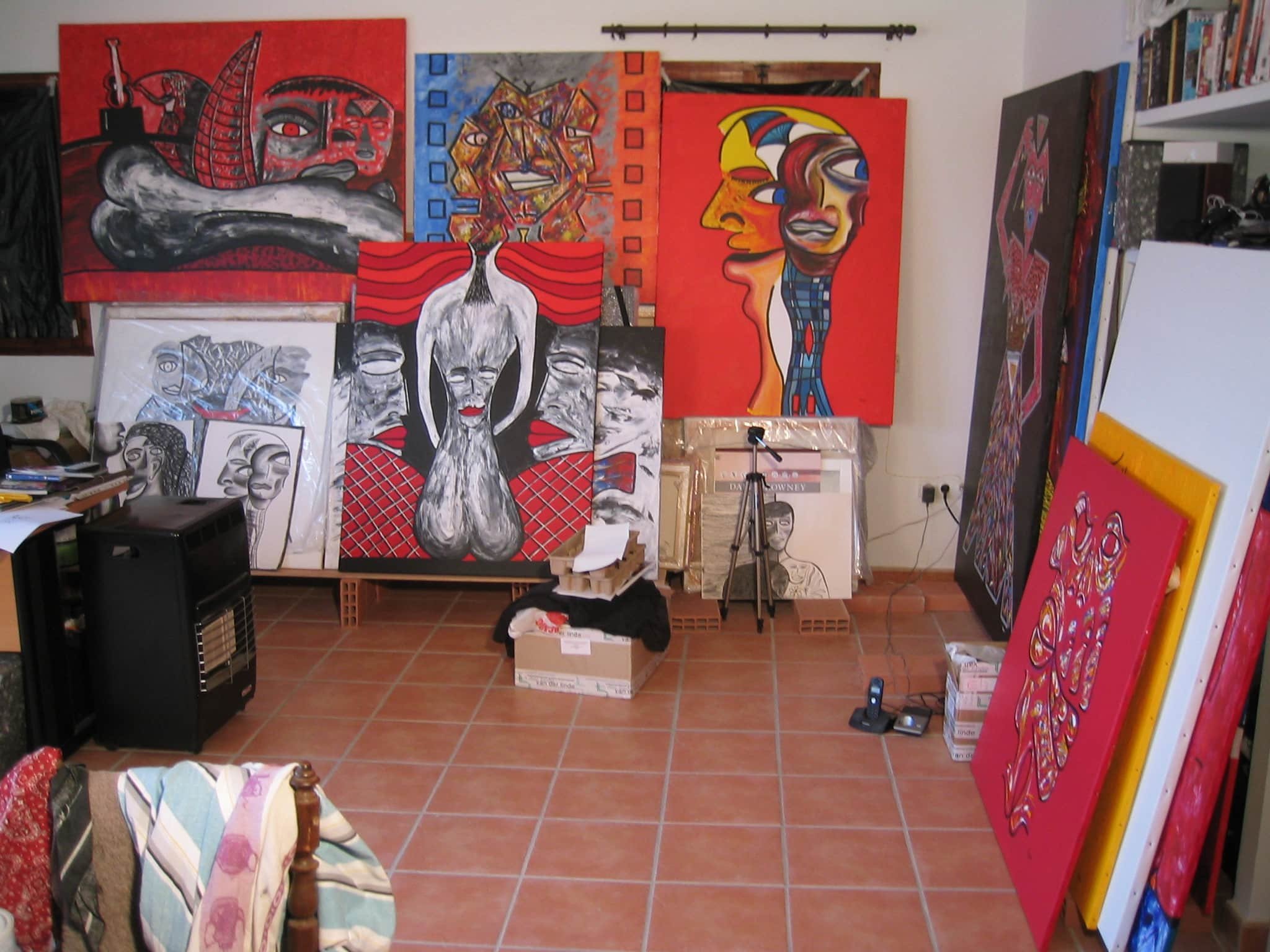 Inside Zarum's art studio in Spain with multiple colorful paintings on the walls and leaning against furniture. Paintings feature abstract and surreal faces and figures.
