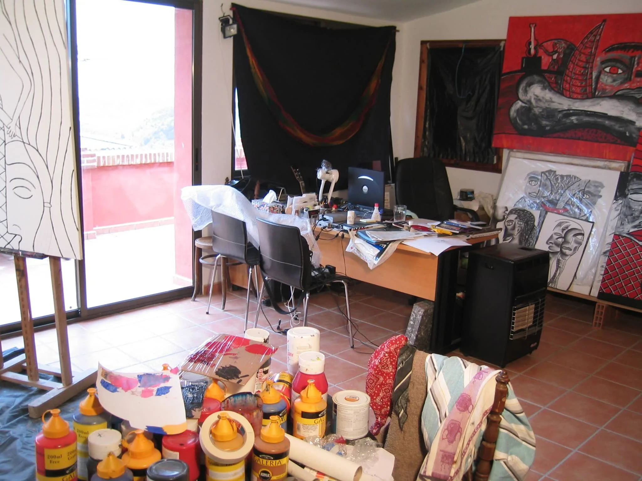 Inside Zarum's art studio in Spain, with paintings, supplies, and clutter. Artworks are leaning against the wall and on the desk, with some featuring abstract faces and designs. The room has a large glass door leading to an outdoor space.
