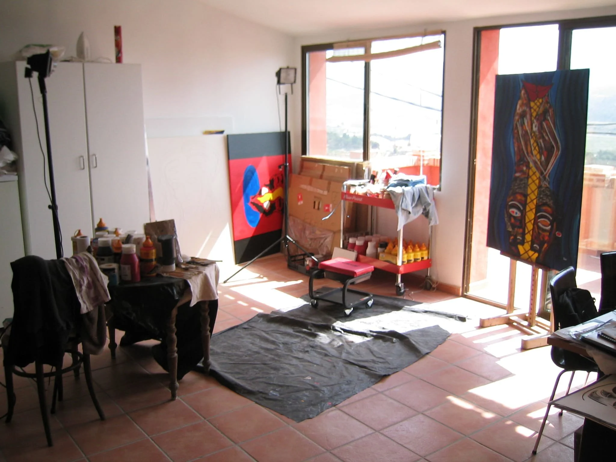 Inside Zarum's art studio in Spain. with paintings, art supplies, and a workspace by large windows on a tiled floor.