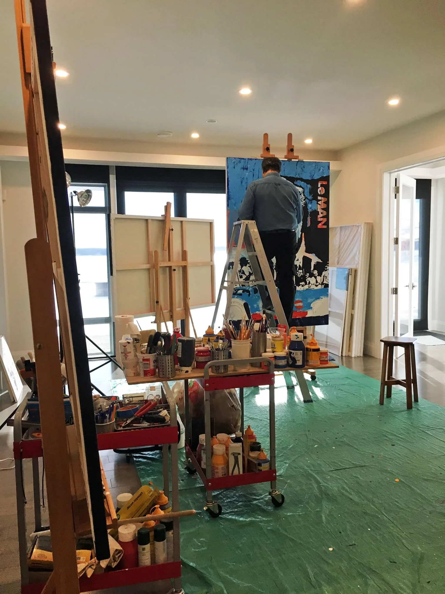  The artist Zarum in his studio in Canada painting a large canvas on an easel while standing on a ladder in a well-lit room. 