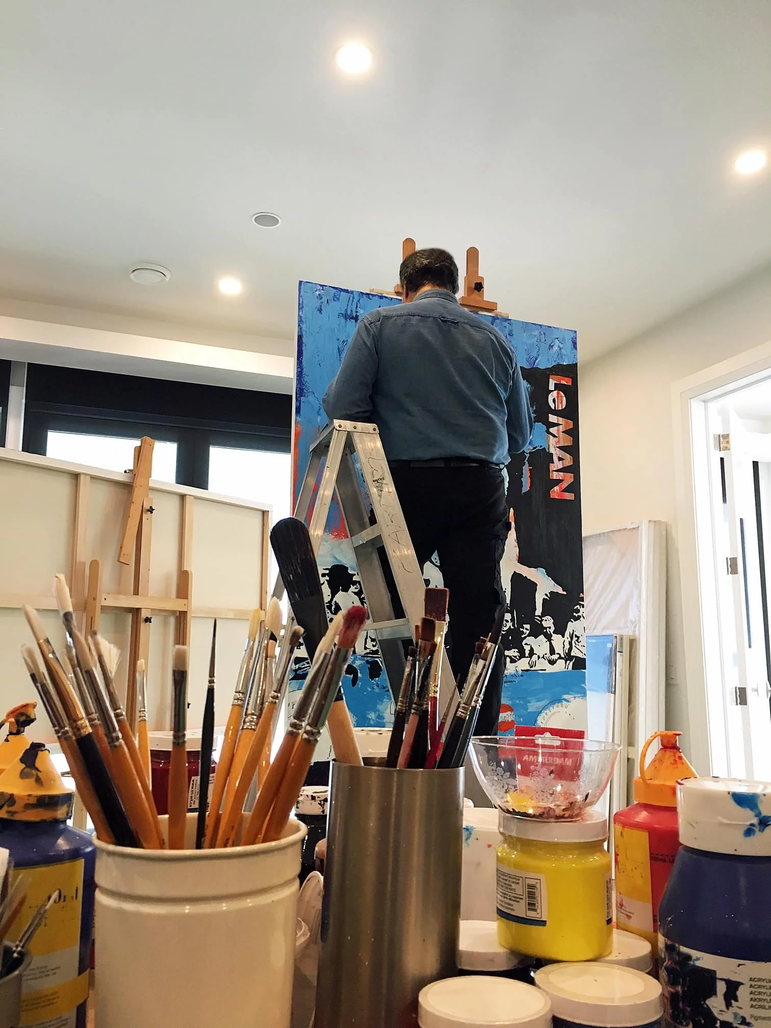 The artist Zarum painting a large canvas with a colorful, abstract scene in a well-lit studio, surrounded by painting supplies in the foreground.