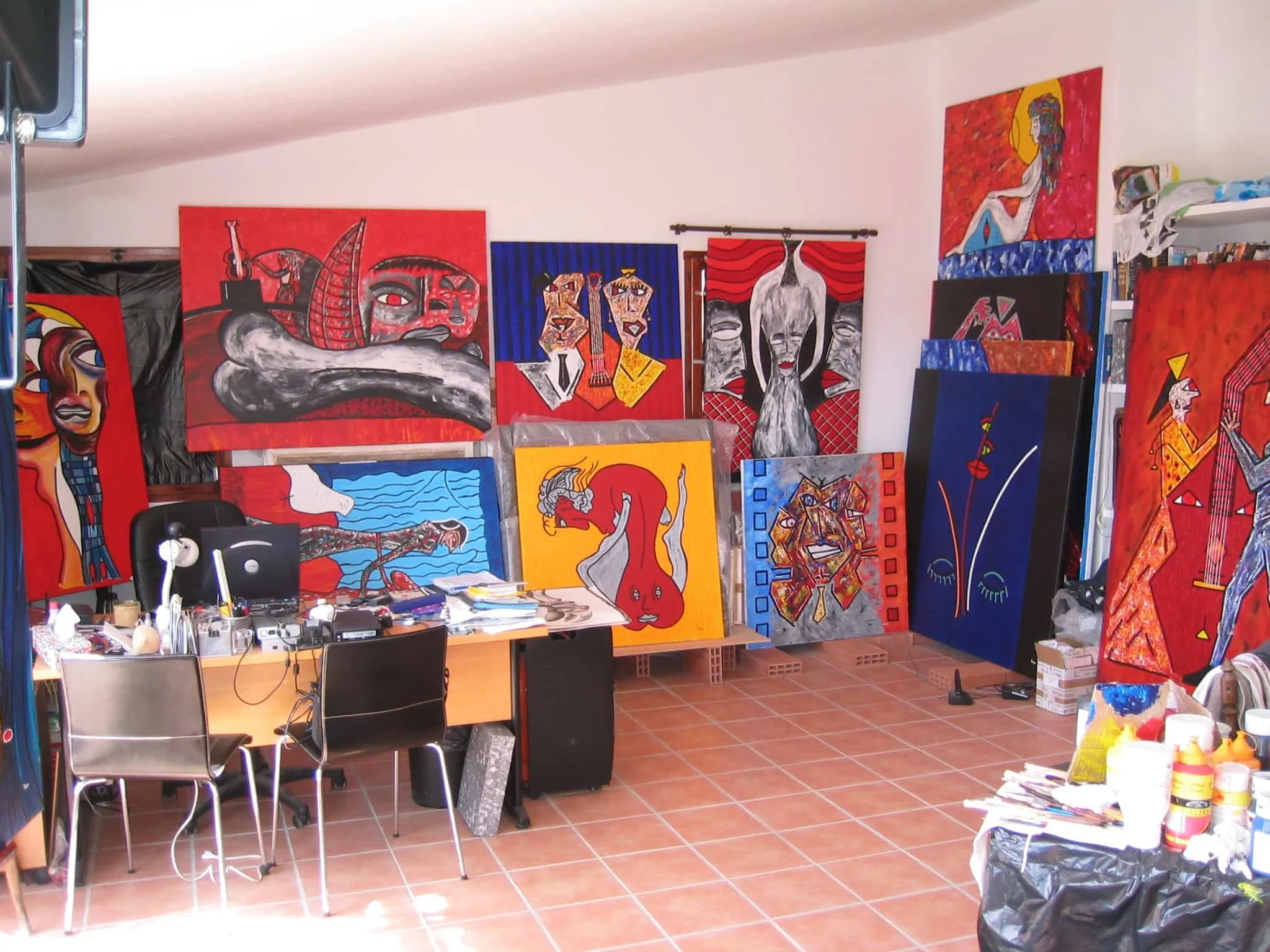 Inside Zarum's art studio in Spain with colorful abstract paintings stacked and displayed on the walls and floor, cluttered desk with art supplies and chairs.