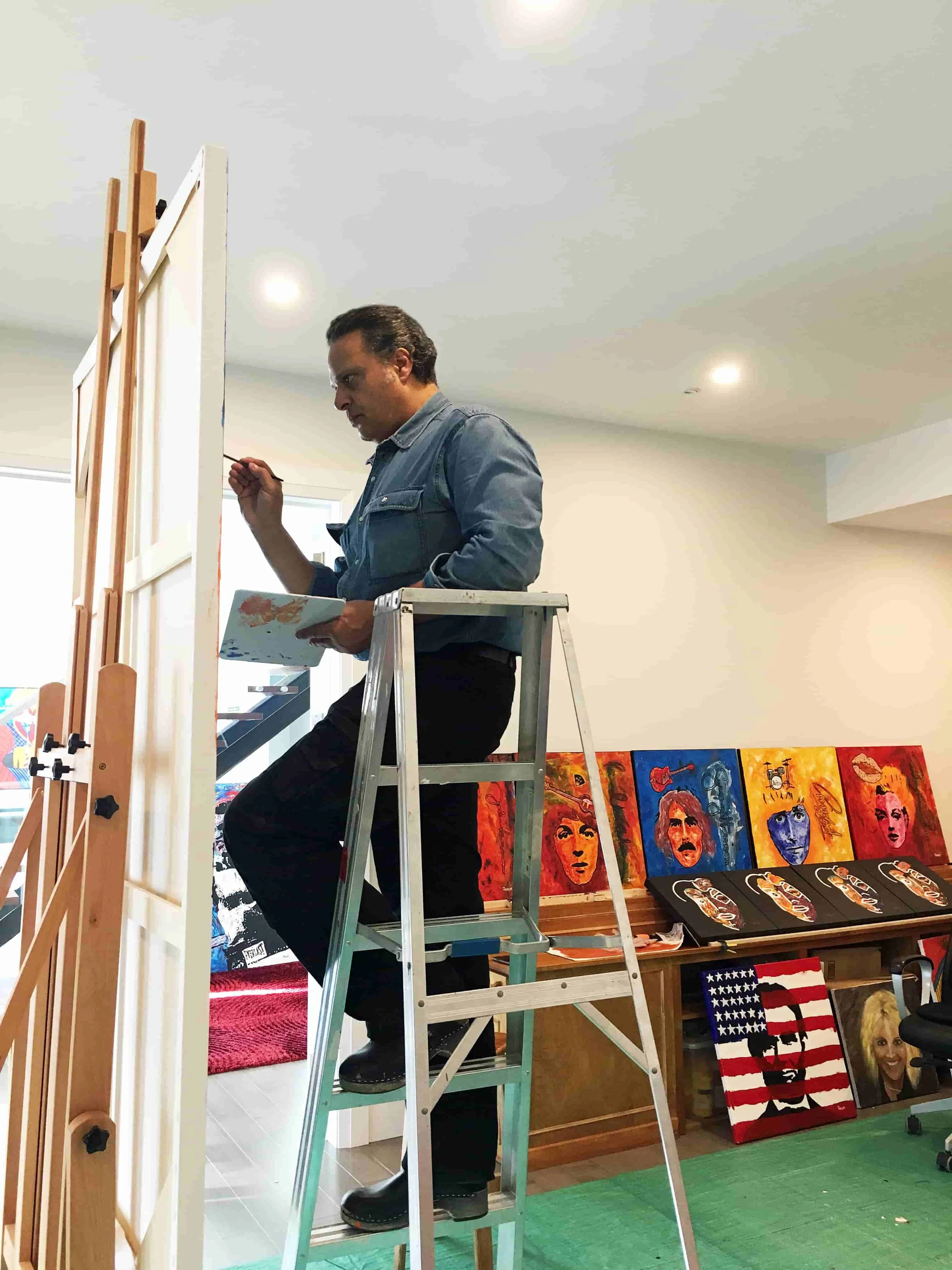 Zarum in his art studio in Canada standing on a ladder painting,, surrounded by colorful paintings and artwork on display.