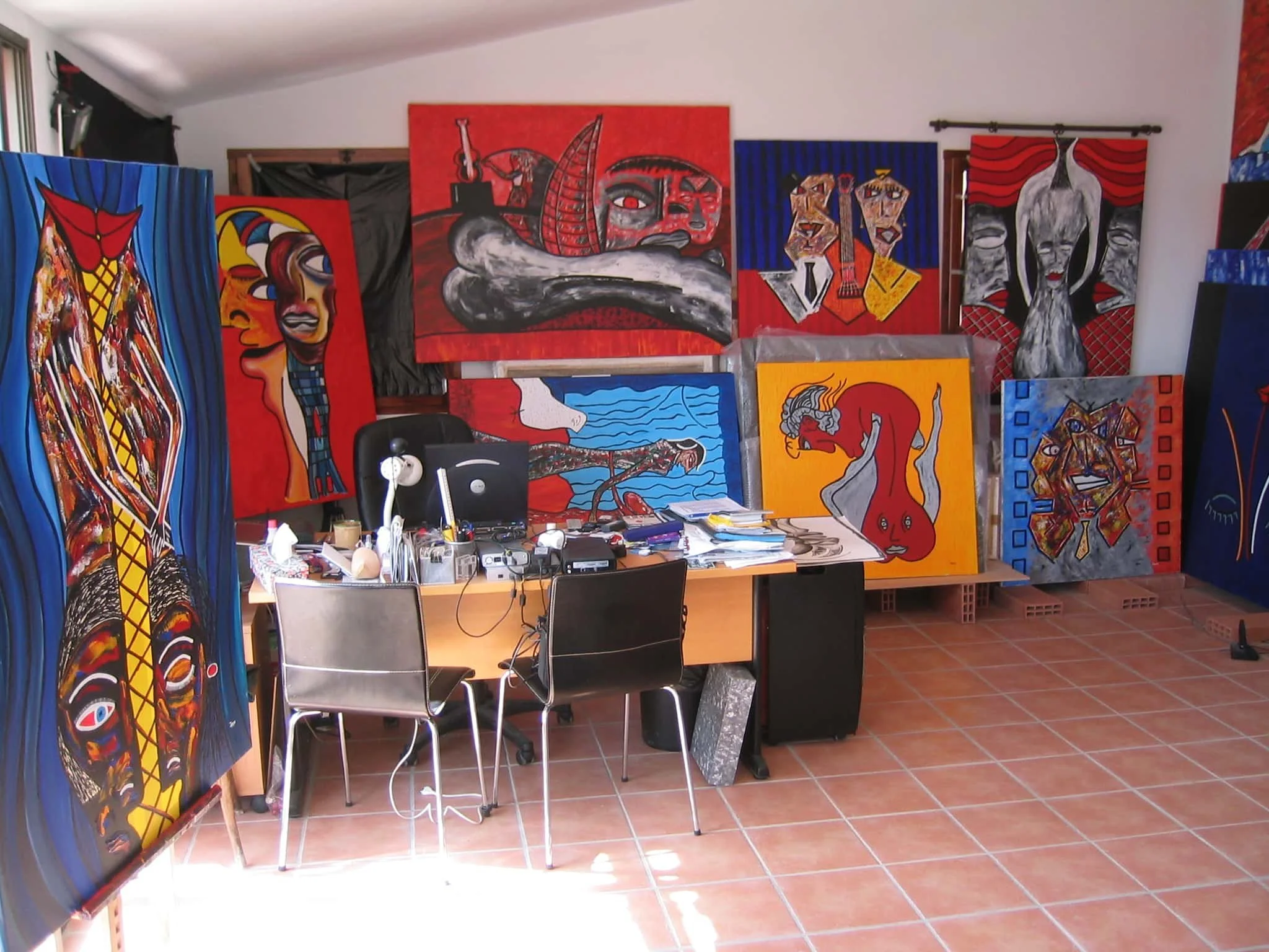 Inside Zarum's art studio in Spain with various colorful abstract paintings on the walls and leaning against the walls, and a cluttered desk with papers and art supplies.