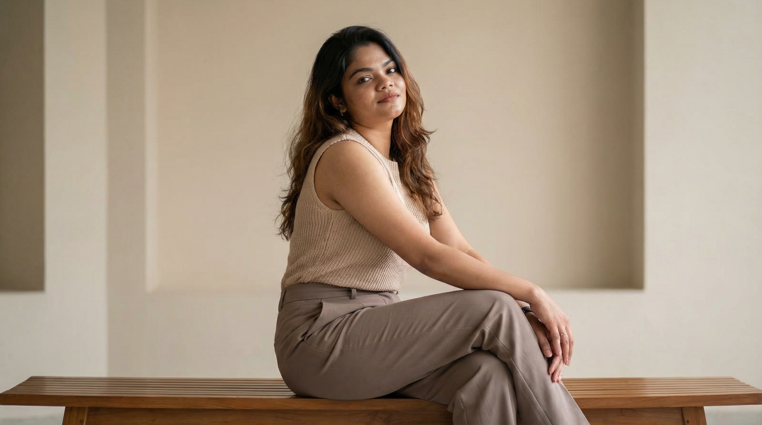 A woman with black and brown wavy hair, wearing a beige sleeveless top and tan pants, sitting on a wooden bench with a simple background.