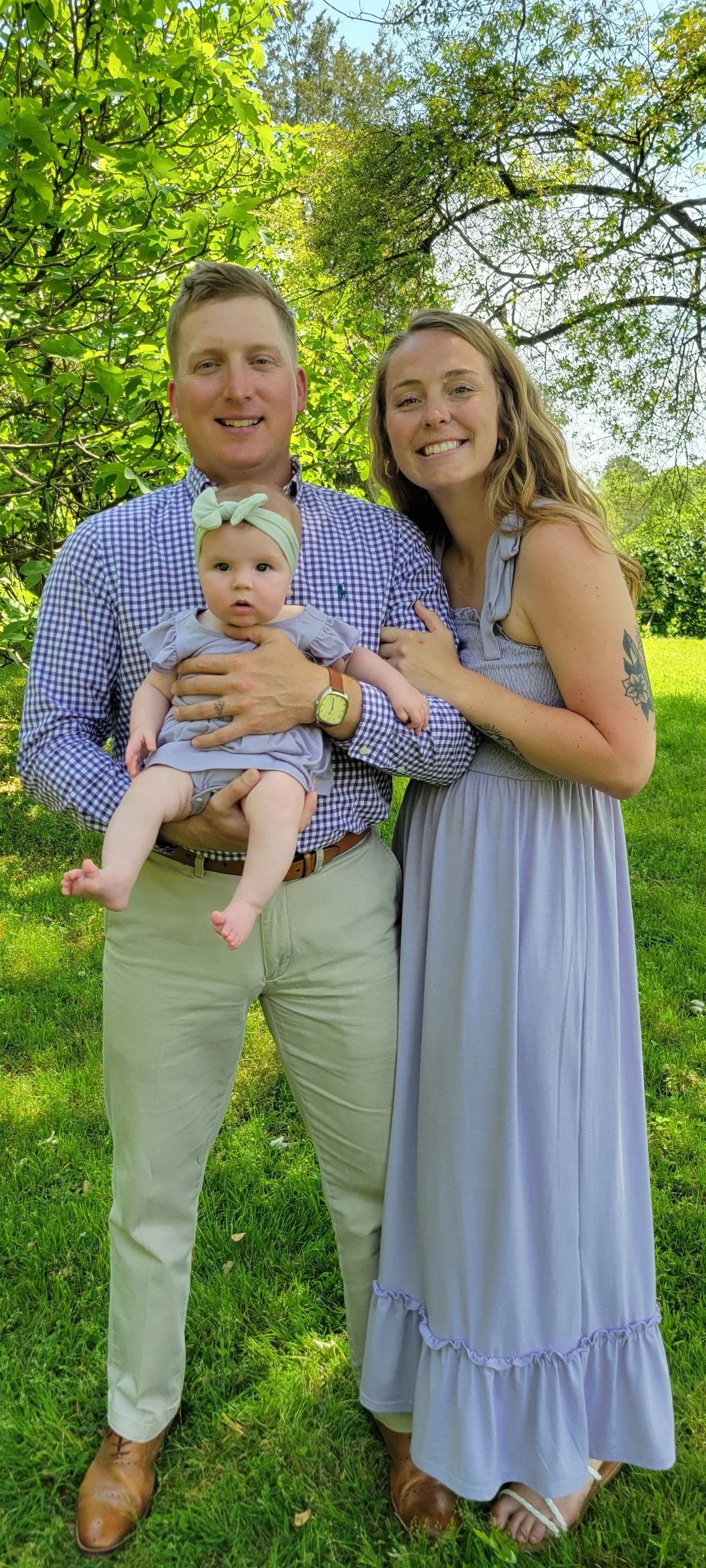 A family of three outdoors on a sunny day with green trees and grass in the background. The father is holding a baby girl dressed in a gray outfit and headband. The mother, wearing a light gray dress, is standing beside them, all smiling at the camera.