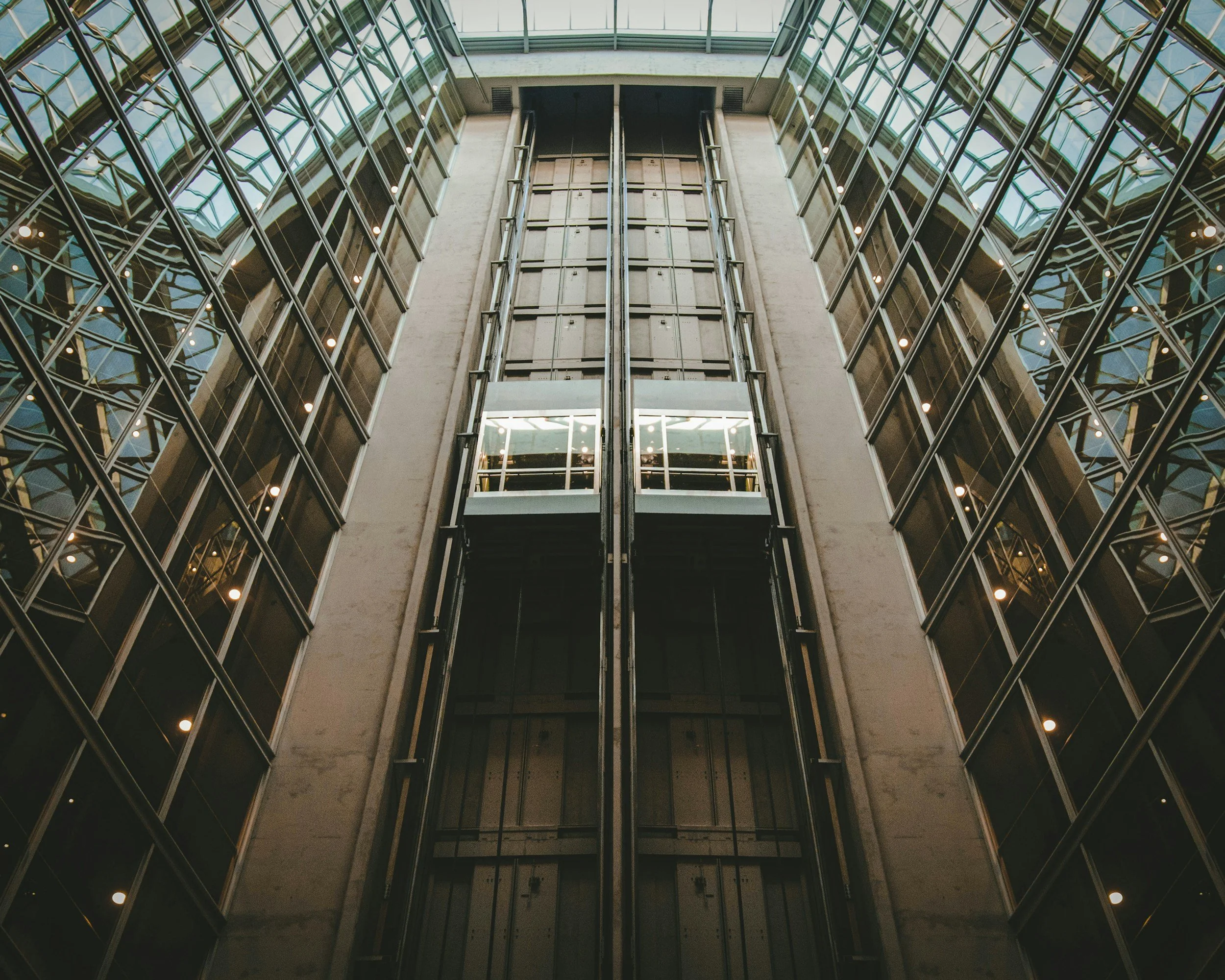 Interior view of a modern multi-story building with glass walls and an elevator shaft in the center, looking upward from the ground floor.