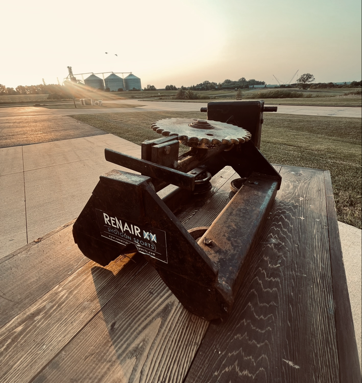 A metal shooting bench with a gear mechanism on a wooden surface outdoors, with a rural landscape including silos and fields in the background during sunset.