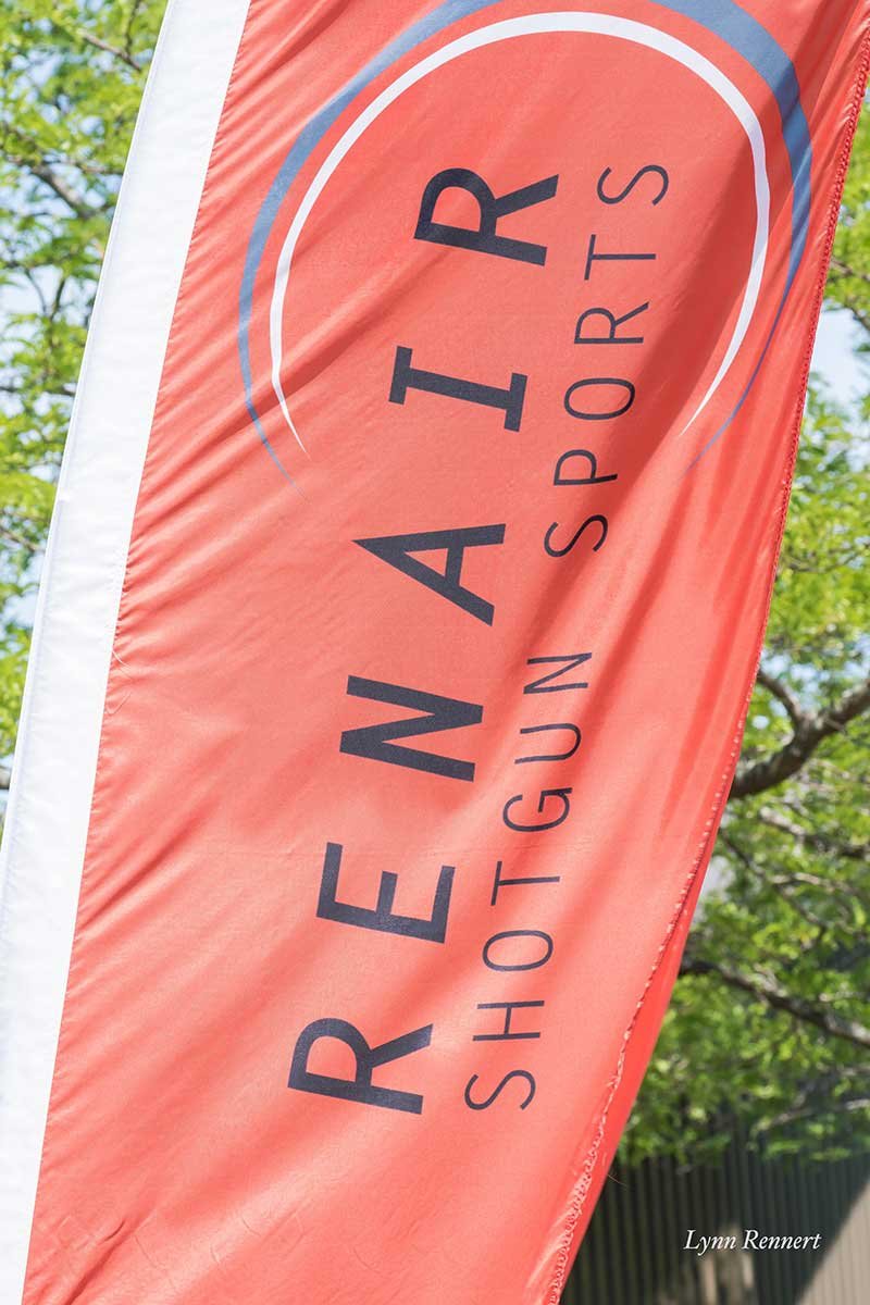 A red and white flag with the words 'RENAIR SHOTS' written on it, partially seen, with green trees and a building in the background.