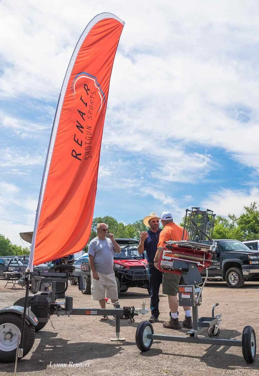 Three men talking outdoors at an event with a large orange flag reading "ReNAT Shootgun Sports" and a firearm carrier stand with shotguns in the foreground. Several vehicles are parked in the background.