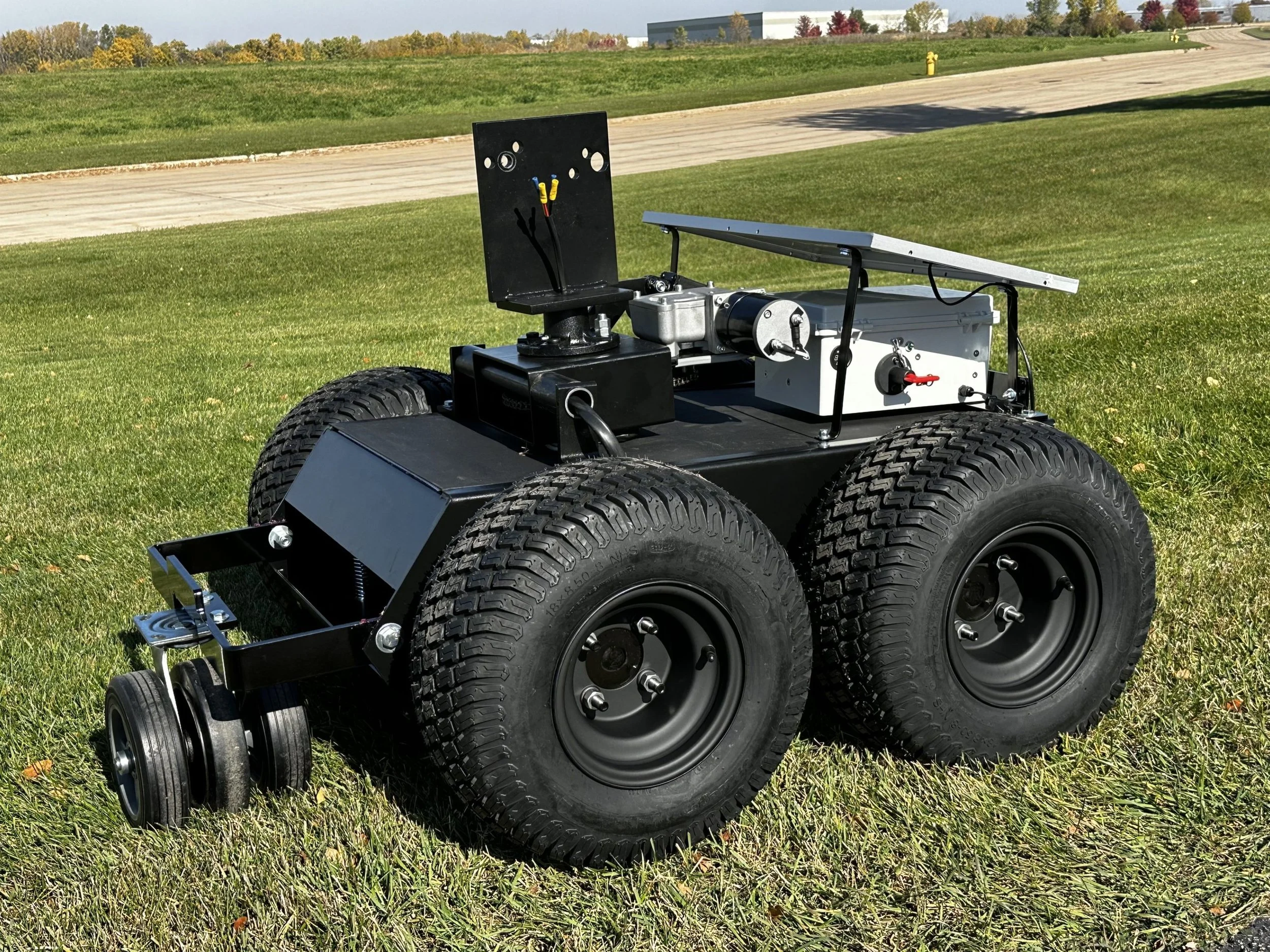 A four-wheeled robotic vehicle with large off-road tires, a black and silver body, and electronic components mounted on top, situated on a grassy field with a road and industrial buildings in the background.