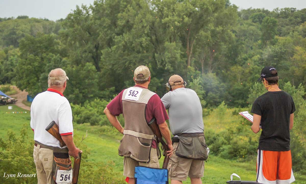 Four people at a shooting range with guns, targets, and greenery in the background.