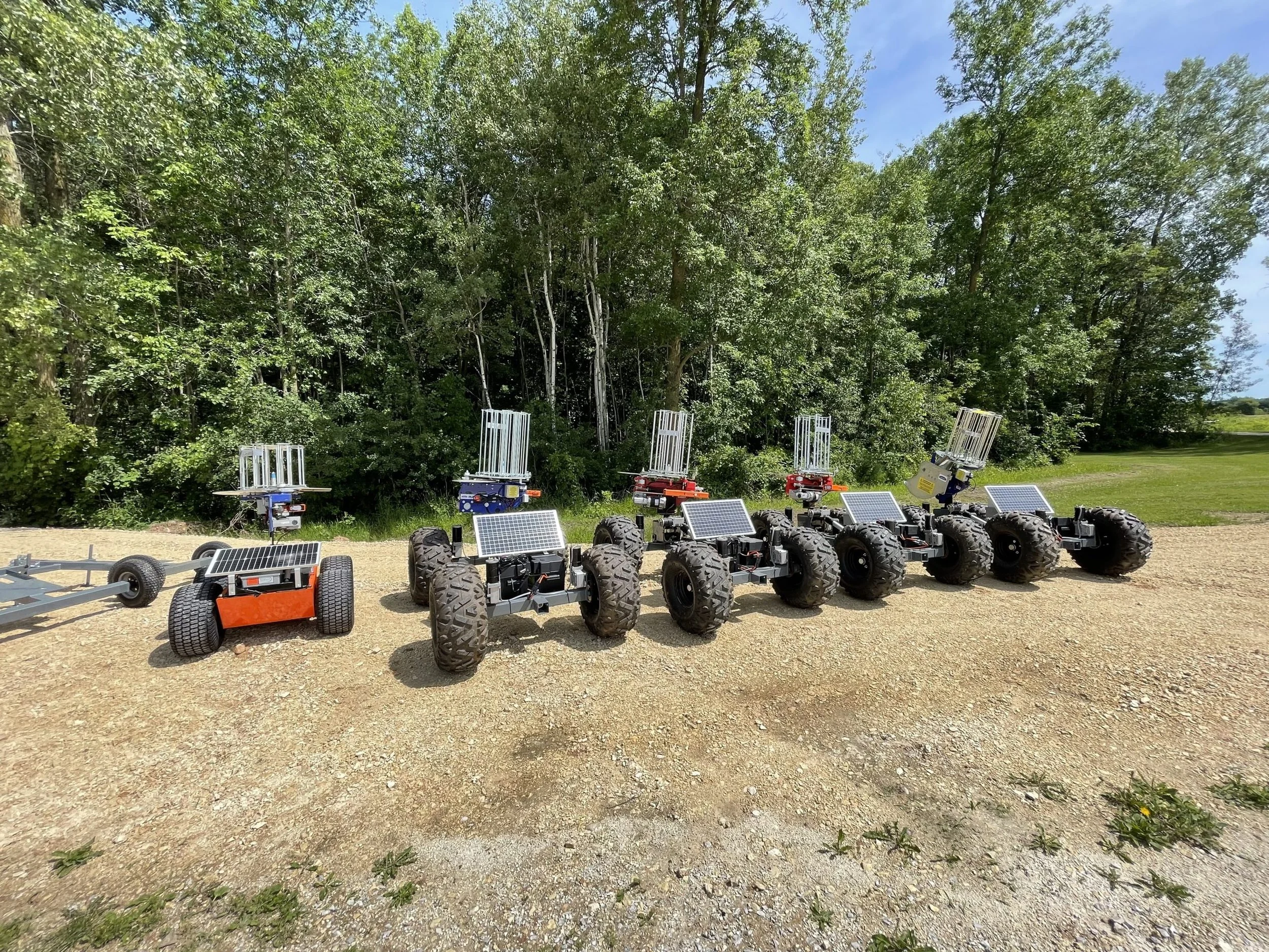 Multiple small robotic vehicles with solar panels and antennas are lined up outdoors on a dirt surface with green trees and grass in the background.