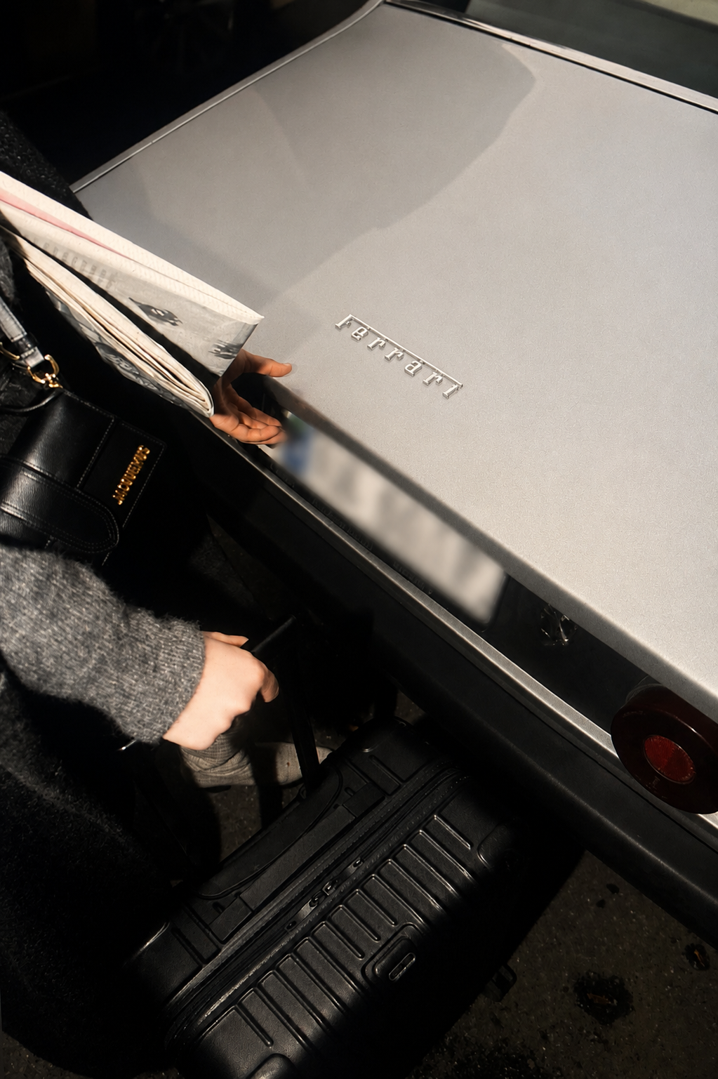 Atelier Onyx - Person greift nach einem Koffer, während sie einen großen Ferrari Karton auf einem Gepäckwagen in einem Flughafen an der Gepäckausgabe hält.