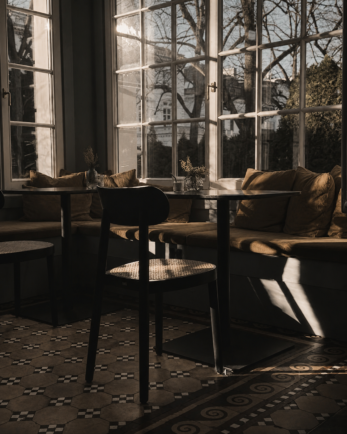 Atelier Onyx - Einen gemütlichen Sitzbereich mit gepolsterten Bänken und Stühlen vor großen Fenster, durch die Sonnenlicht auf den Boden fällt. Draußen erkennt man Bäume und Nachbargebäude.