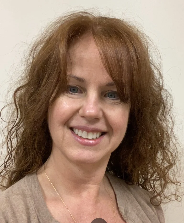 A woman with curly red hair smiling against a plain light-colored background.