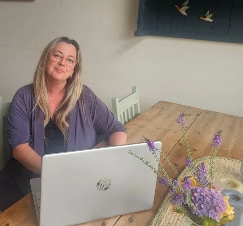 Woman sitting at a dining table with a laptop open in front of her, a basket of purple flowers, and a wall hanging with flying birds in the background.