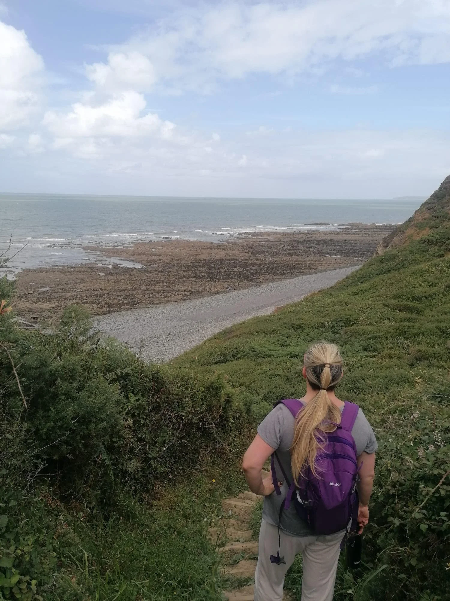 A woman with long blond hair, wearing a gray shirt and purple backpack, walking down a trail through green foliage toward a coastal view of rocky shoreline and the ocean with cloudy sky.