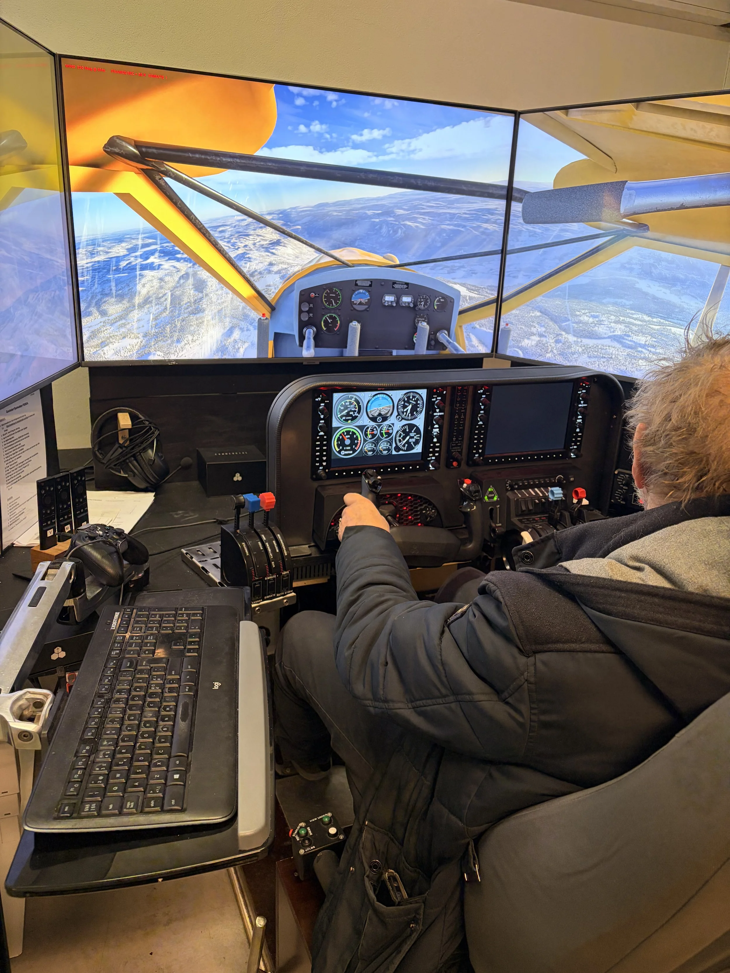 A person in a cockpit with multiple screens and controls, flying over snowy mountains, with a view of the sky and clouds outside.