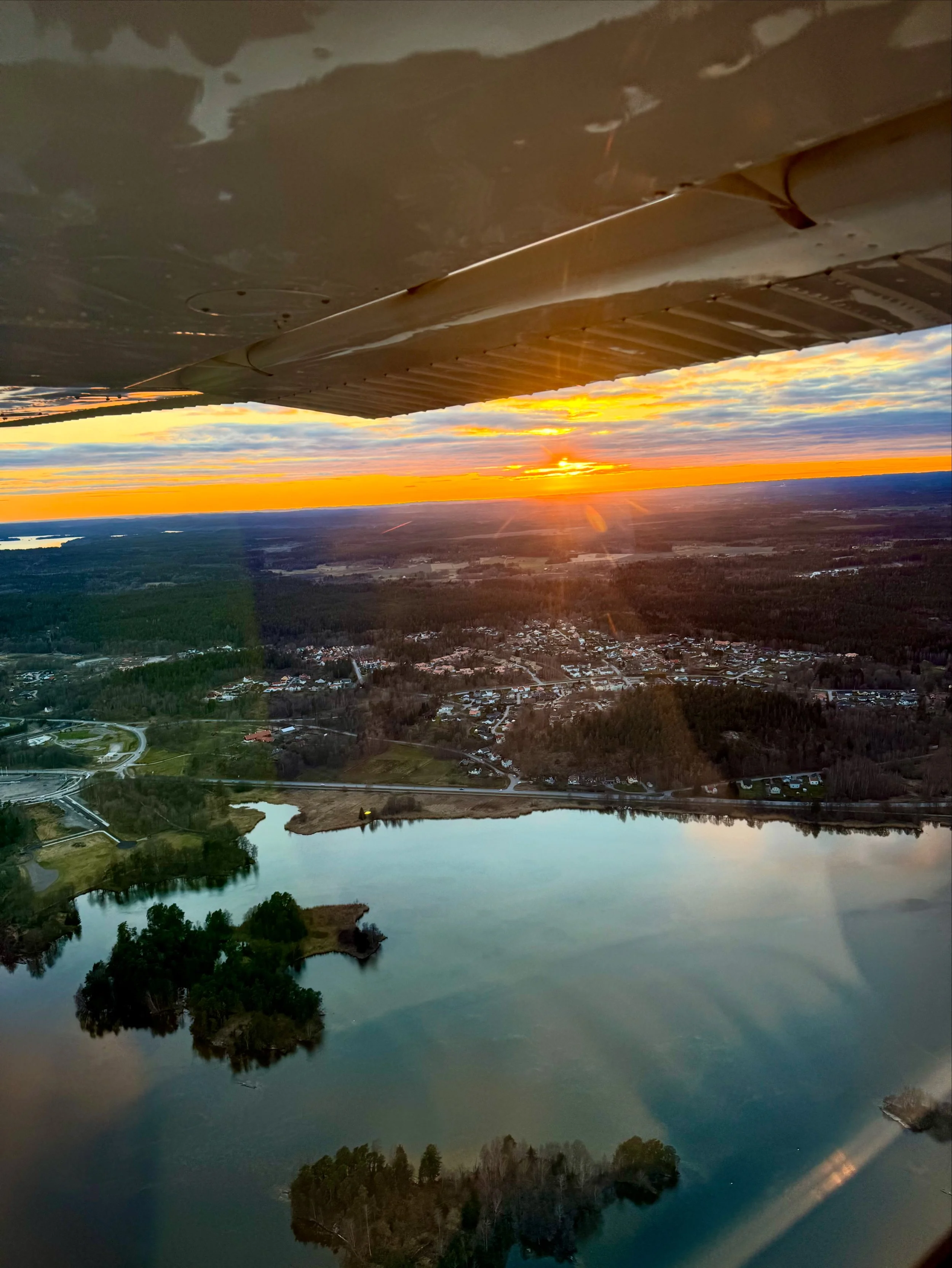 Aerial view of a lake with small islands, residential area, and forested land during sunset with the wing of an aircraft visible in the sky.