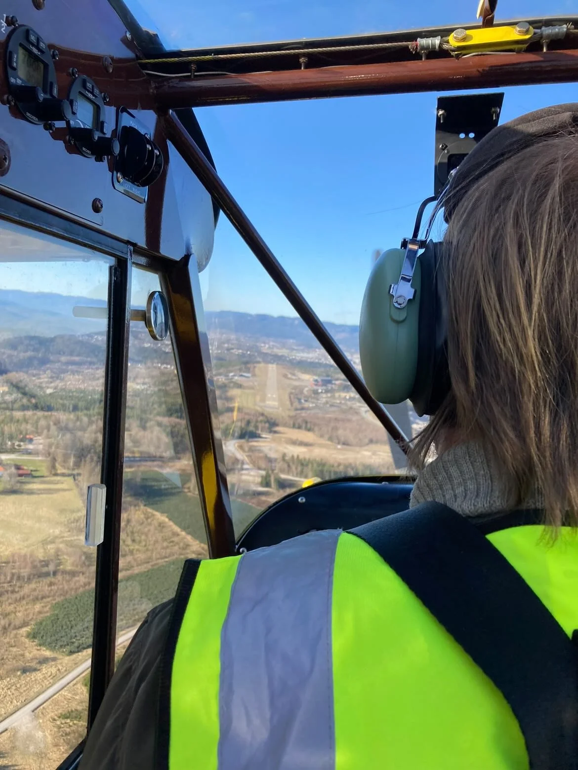 A person wearing a high-visibility safety vest and headphones flying in a small aircraft over a rural landscape with fields and roads on a clear day.