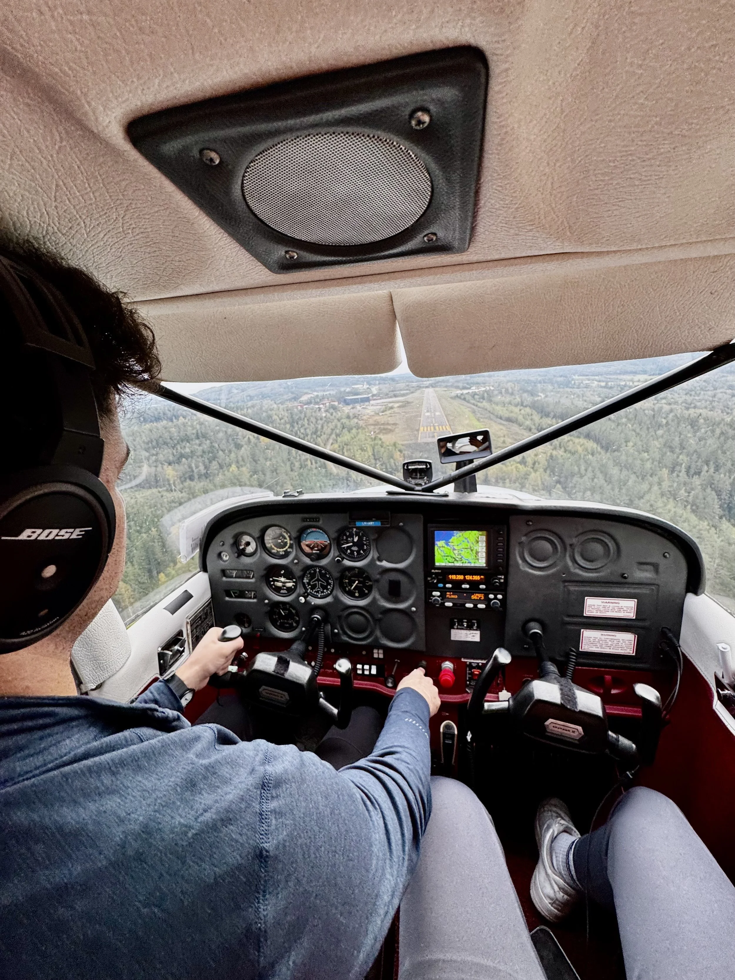 Person wearing Bose headphones and a denim jacket flying a small aircraft, with a view of a runway and green landscape ahead.